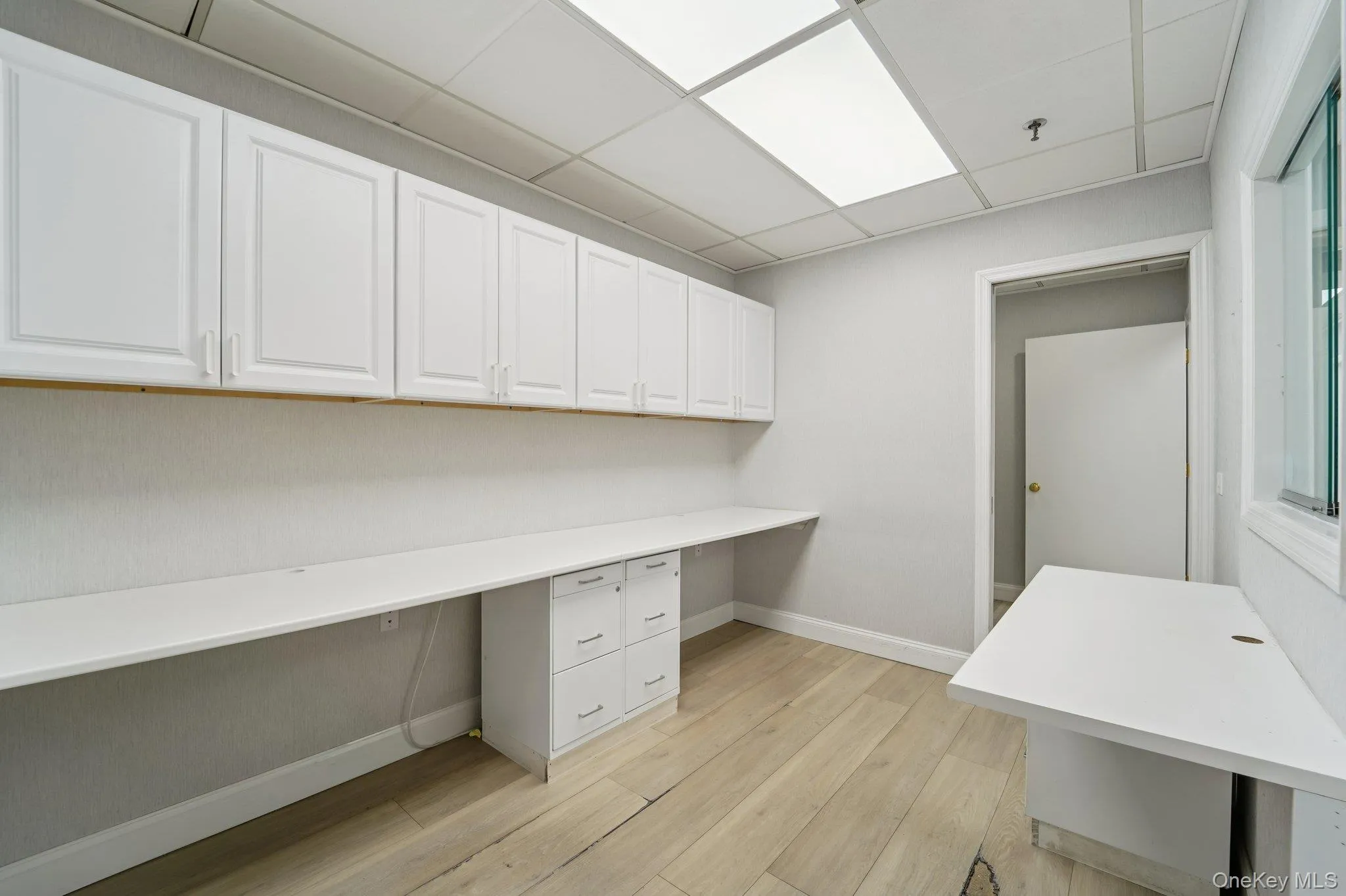 Unfurnished office with a drop ceiling, built in desk, and light wood-style flooring Unfurnished office with a drop ceiling, built in desk, and light wood-style flooring