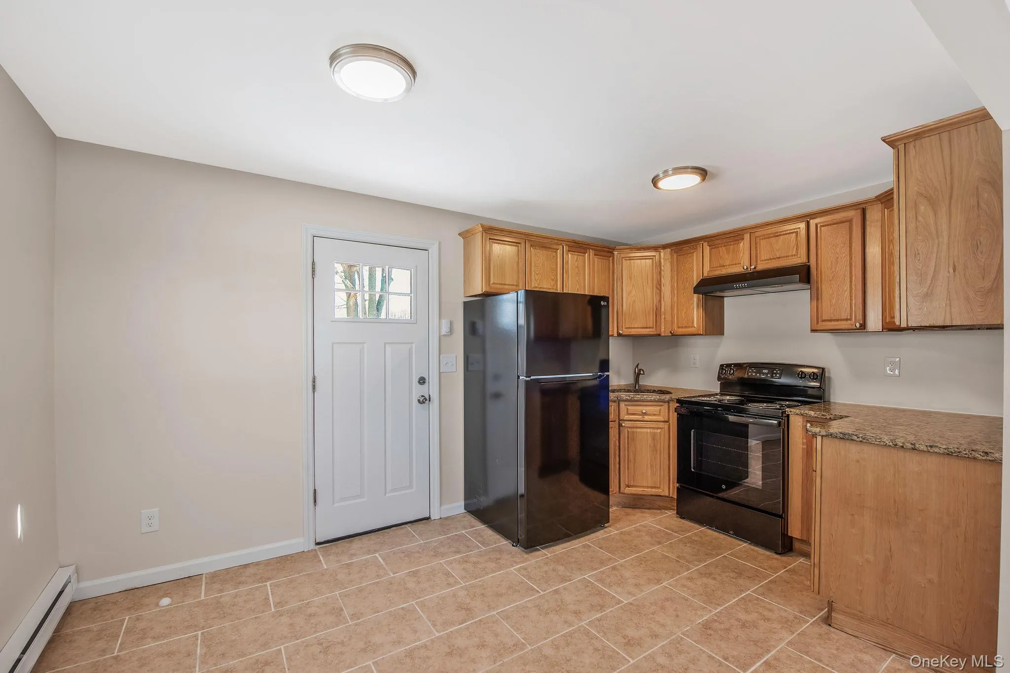 Kitchen with black appliances, baseboard heating, dark stone countertops, under cabinet range hood, and light tile patterned floors Kitchen with black appliances, baseboard heating, dark stone countertops, under cabinet range hood, and light tile patterned floors