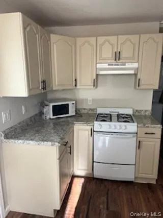 Kitchen with white appliances, under cabinet range hood, dark wood-style flooring, light stone counters, and cream cabinets Kitchen with white appliances, under cabinet range hood, dark wood-style flooring, light stone counters, and cream cabinets