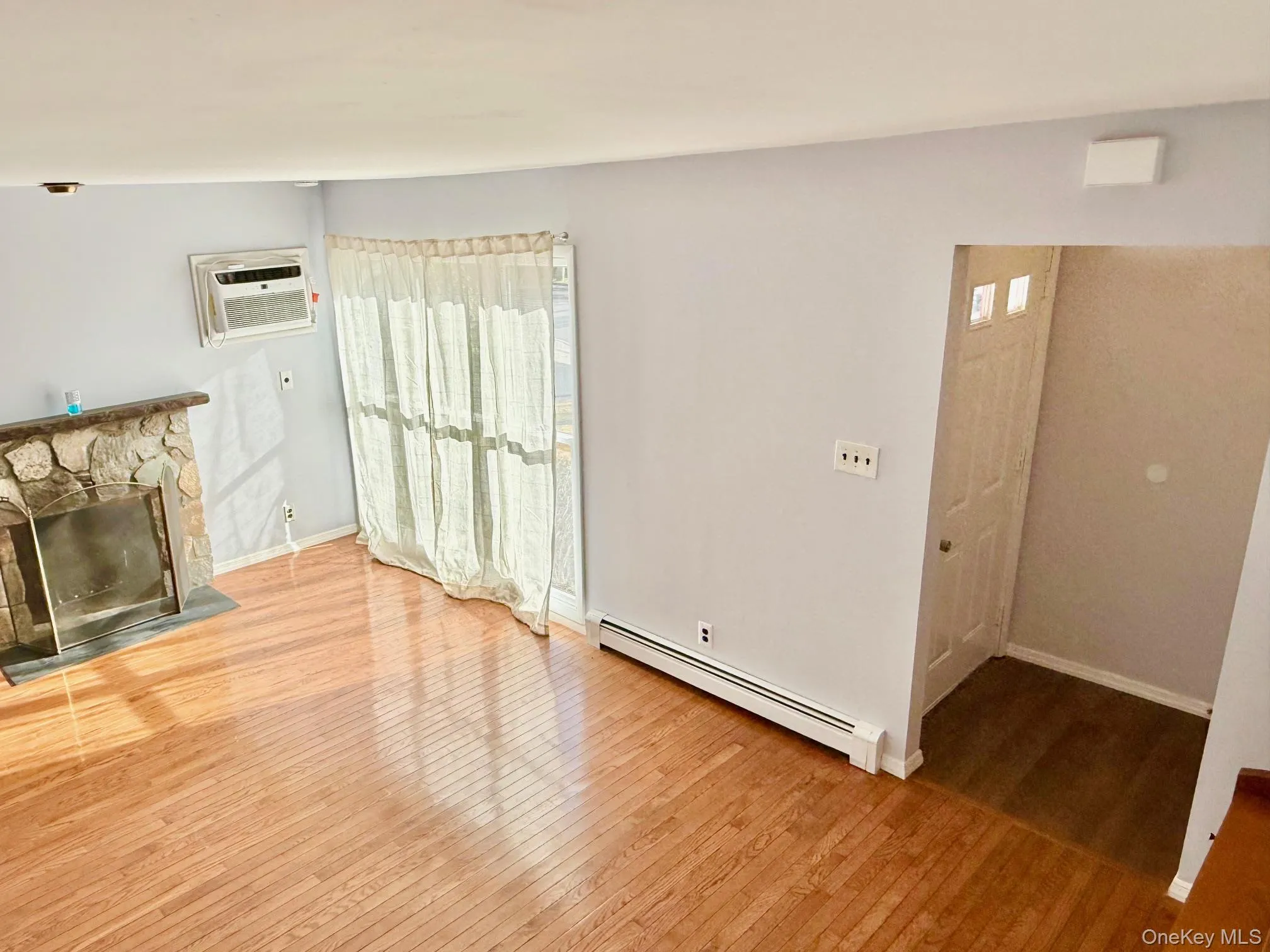 Unfurnished living room featuring a baseboard heating unit, light wood finished floors, a fireplace, and a wall mounted AC Unfurnished living room featuring a baseboard heating unit, light wood finished floors, a fireplace, and a wall mounted AC