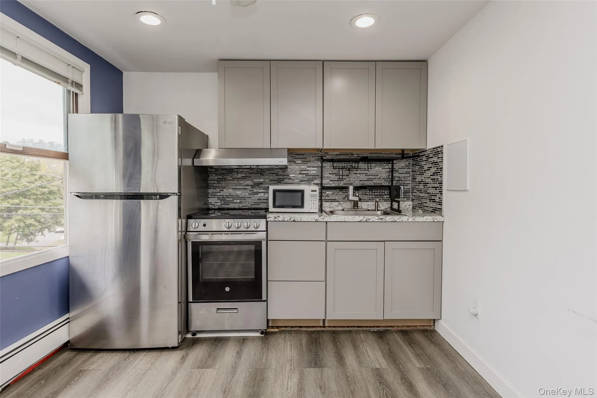 Kitchen featuring gray cabinetry, appliances with stainless steel finishes, a baseboard radiator, backsplash, and ventilation hood Kitchen featuring gray cabinetry, appliances with stainless steel finishes, a baseboard radiator, backsplash, and ventilation hood