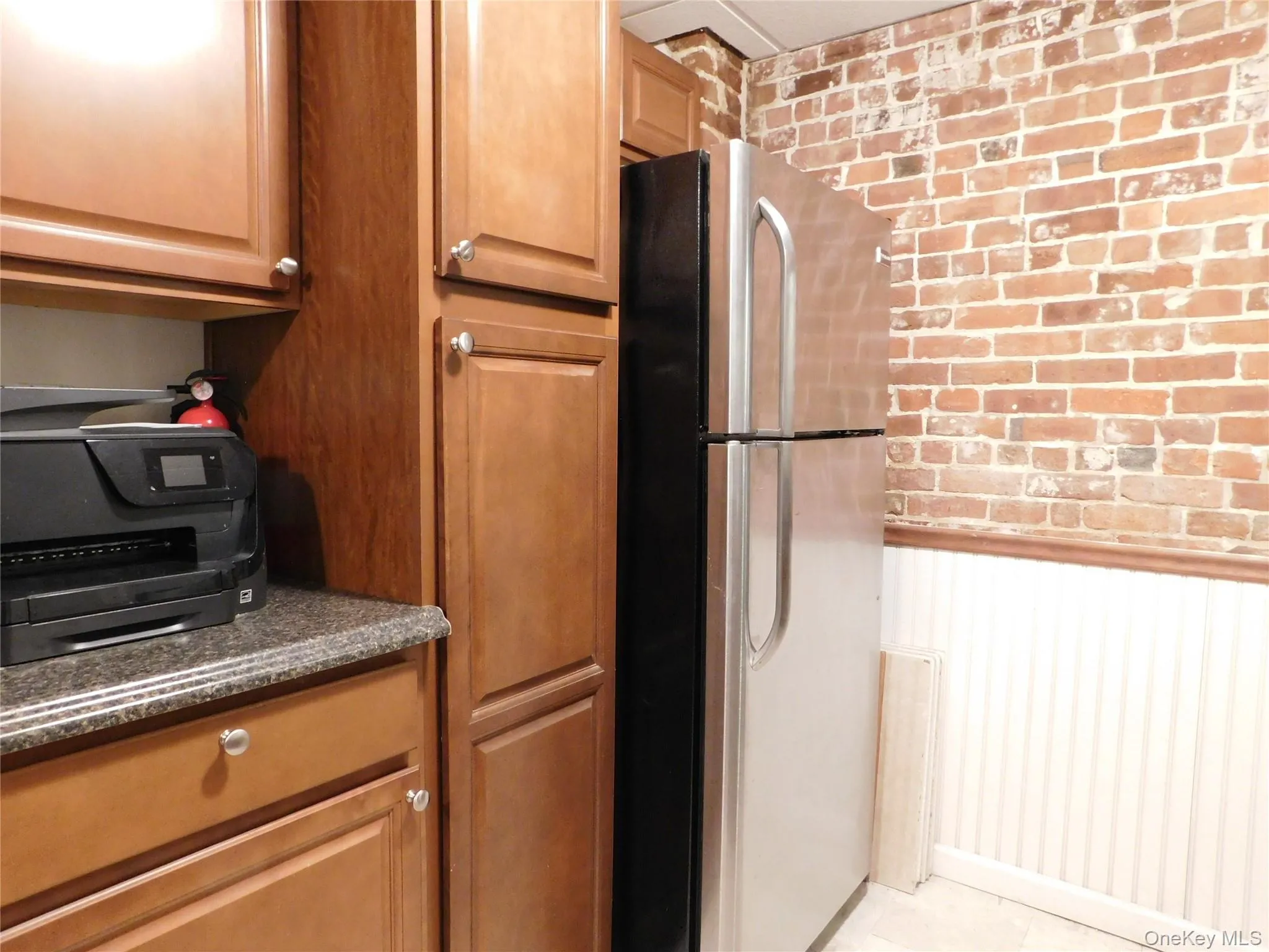Kitchen with brick wall, freestanding refrigerator, brown cabinetry, dark stone countertops, and a wainscoted wall Kitchen with brick wall, freestanding refrigerator, brown cabinetry, dark stone countertops, and a wainscoted wall