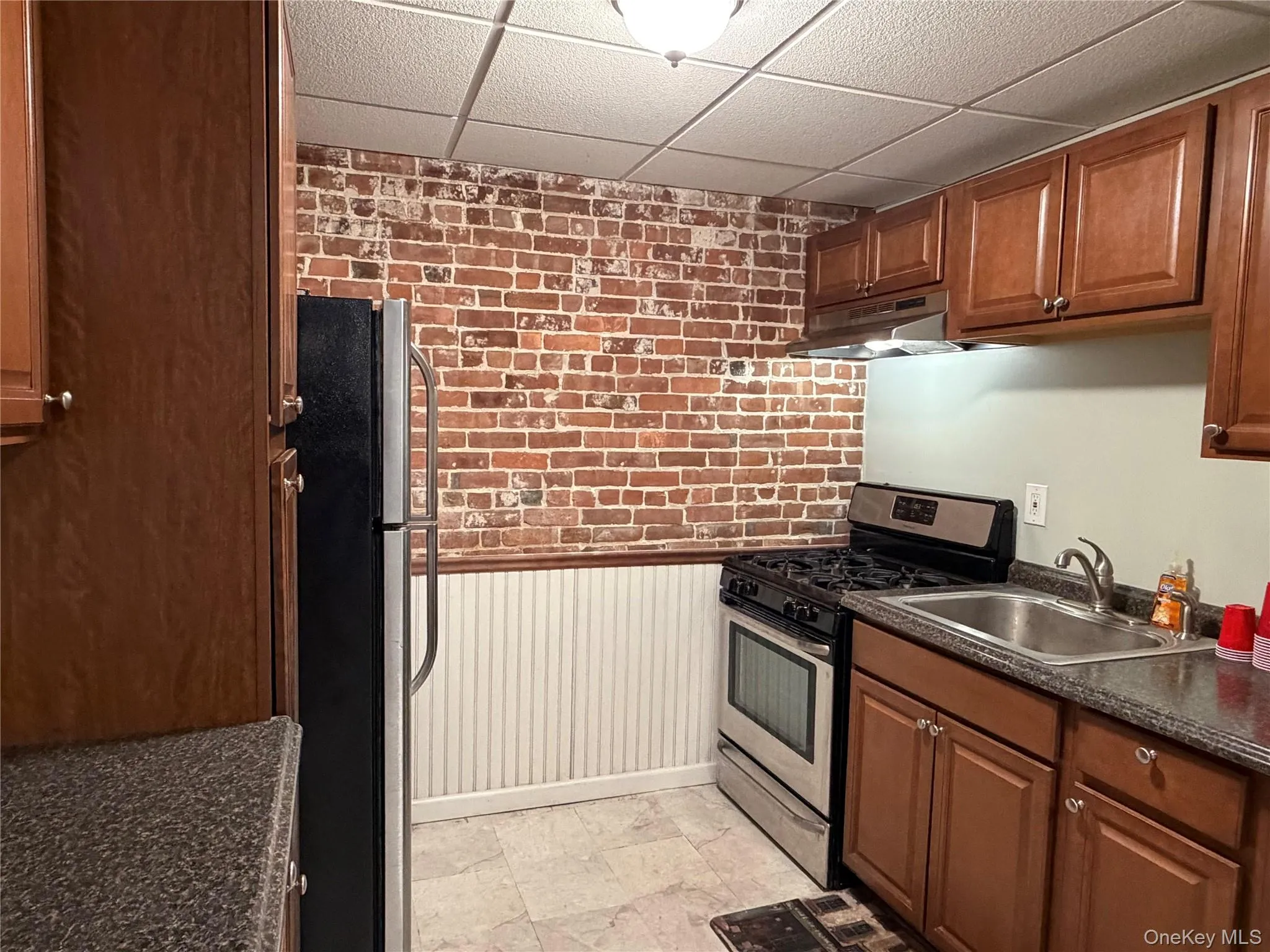 Kitchen featuring brick wall, stainless steel appliances, a paneled ceiling, dark countertops, and brown cabinets Kitchen featuring brick wall, stainless steel appliances, a paneled ceiling, dark countertops, and brown cabinets