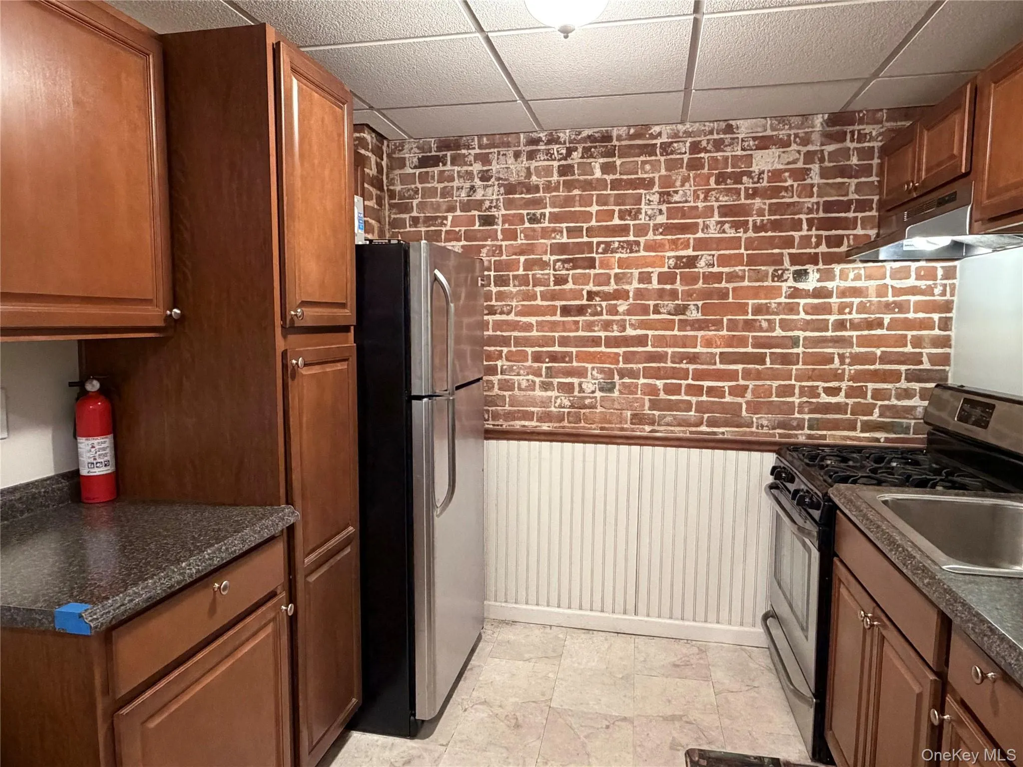 Kitchen featuring brick wall, wainscoting, dark countertops, stainless steel appliances, and a paneled ceiling Kitchen featuring brick wall, wainscoting, dark countertops, stainless steel appliances, and a paneled ceiling