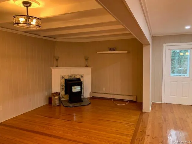 Unfurnished living room featuring a pellet stove, light wood-type flooring, wood walls, baseboard heating, and beam ceiling Unfurnished living room featuring a pellet stove, light wood-type flooring, wood walls, baseboard heating, and beam ceiling