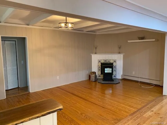 Unfurnished living room featuring beam ceiling, a baseboard heating unit, light wood-style floors, wooden walls, and a pellet stove Unfurnished living room featuring beam ceiling, a baseboard heating unit, light wood-style floors, wooden walls, and a pellet stove