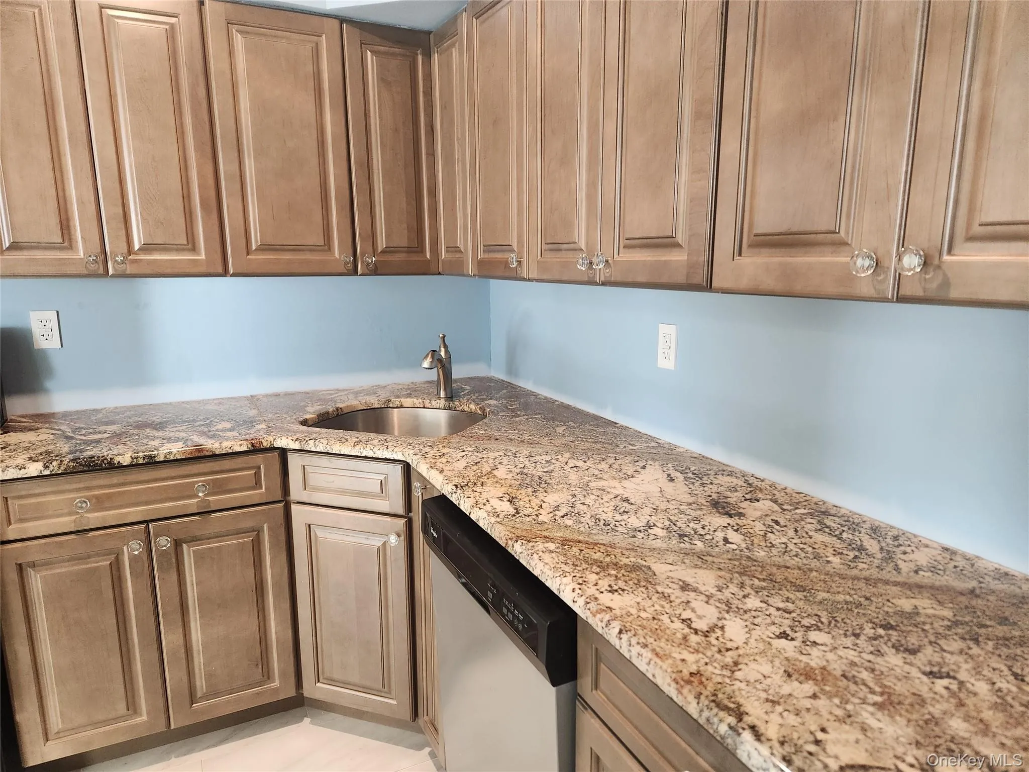 Kitchen featuring light stone countertops, stainless steel dishwasher, and brown cabinetry Kitchen featuring light stone countertops, stainless steel dishwasher, and brown cabinetry