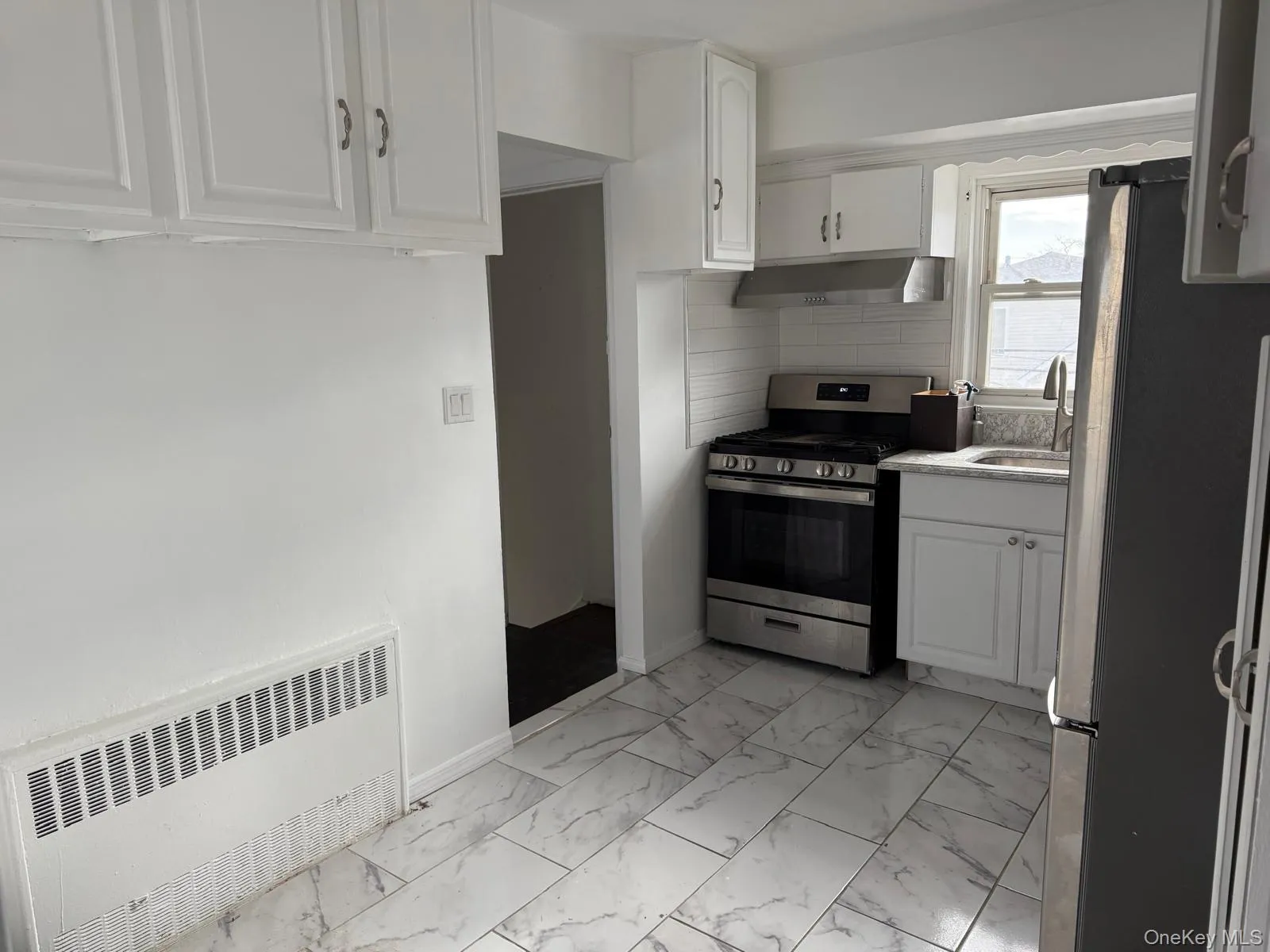 Kitchen with radiator heating unit, white cabinetry, stainless steel appliances, light marble finish flooring, and light countertops Kitchen with radiator heating unit, white cabinetry, stainless steel appliances, light marble finish flooring, and light countertops