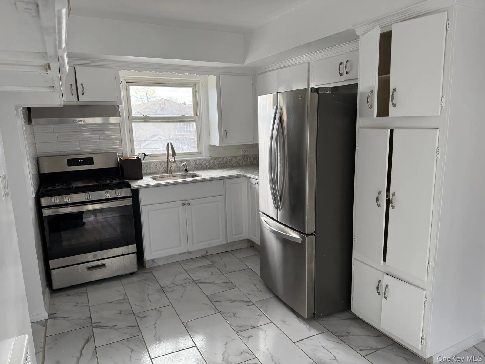 Kitchen with appliances with stainless steel finishes, white cabinetry, light marble finish flooring, and under cabinet range hood Kitchen with appliances with stainless steel finishes, white cabinetry, light marble finish flooring, and under cabinet range hood