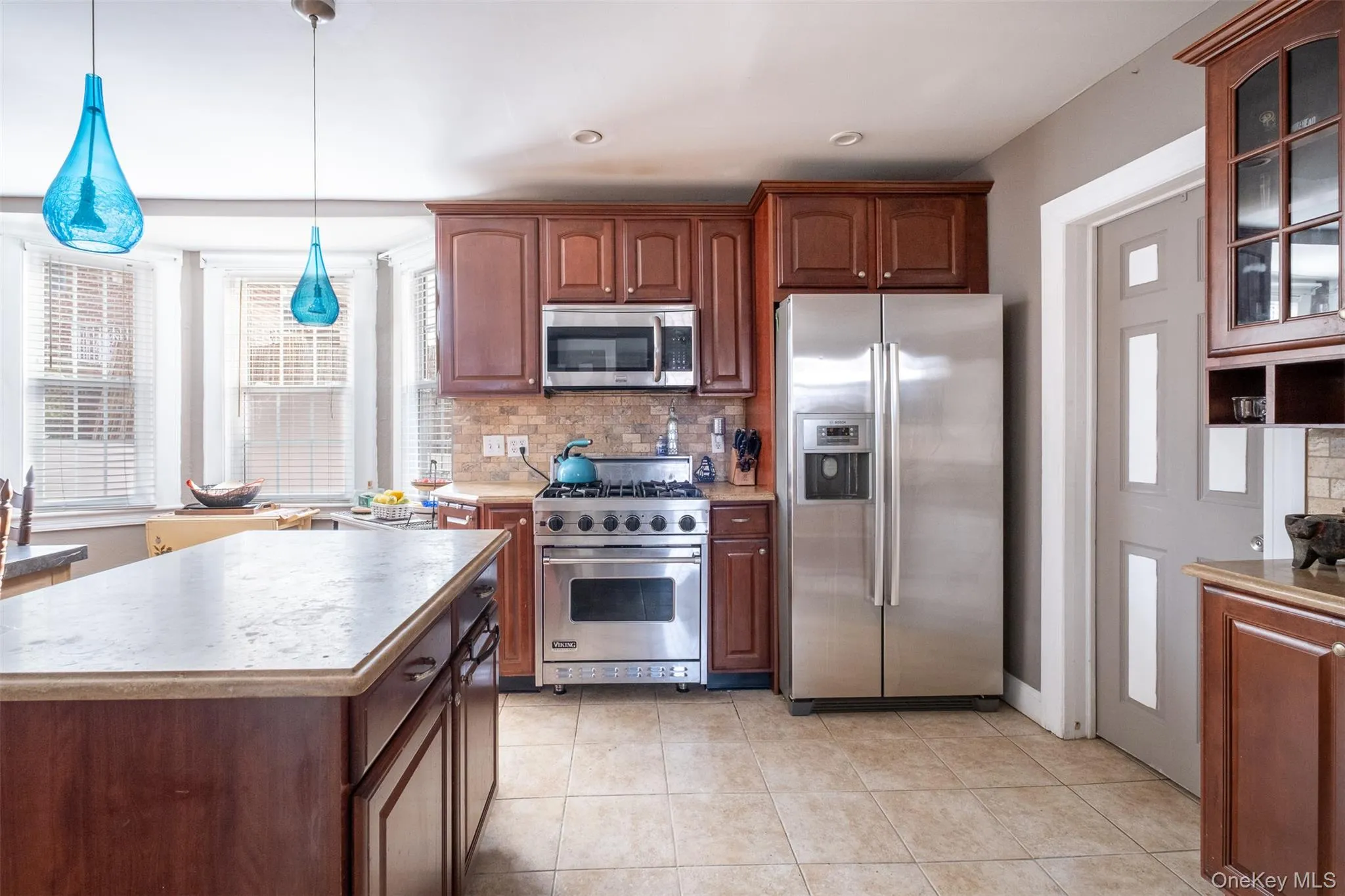 Kitchen with appliances with stainless steel finishes, decorative light fixtures, glass insert cabinets, light countertops, and decorative backsplash Kitchen with appliances with stainless steel finishes, decorative light fixtures, glass insert cabinets, light countertops, and decorative backsplash