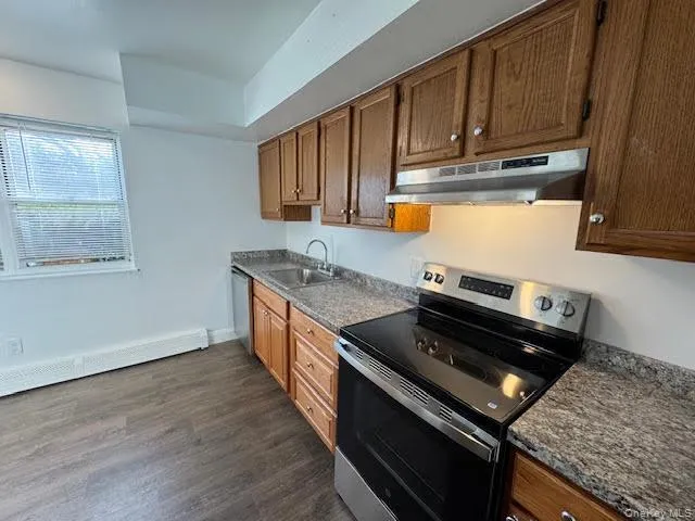 Kitchen with appliances with stainless steel finishes, under cabinet range hood, dark wood-style floors, brown cabinetry, and a baseboard heating unit Kitchen with appliances with stainless steel finishes, under cabinet range hood, dark wood-style floors, brown cabinetry, and a baseboard heating unit