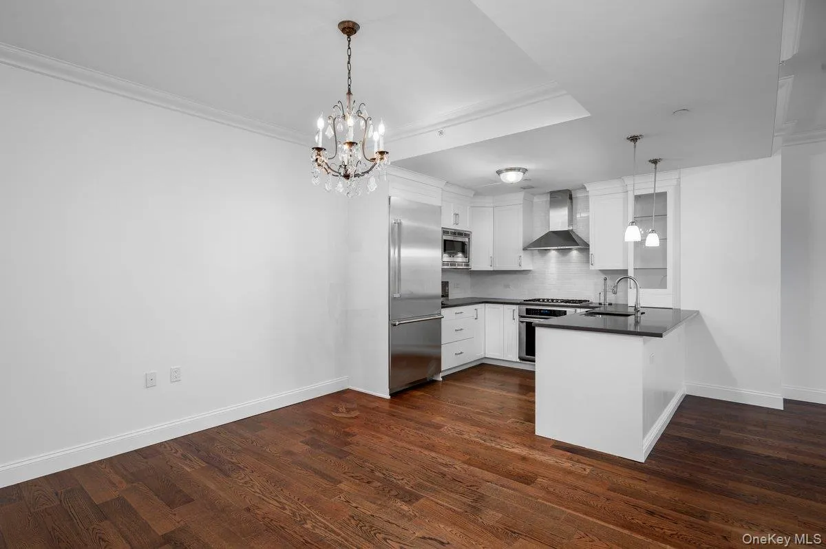 Kitchen featuring white cabinets, a peninsula, wall chimney range hood, dark countertops, and a chandelier Kitchen featuring white cabinets, a peninsula, wall chimney range hood, dark countertops, and a chandelier