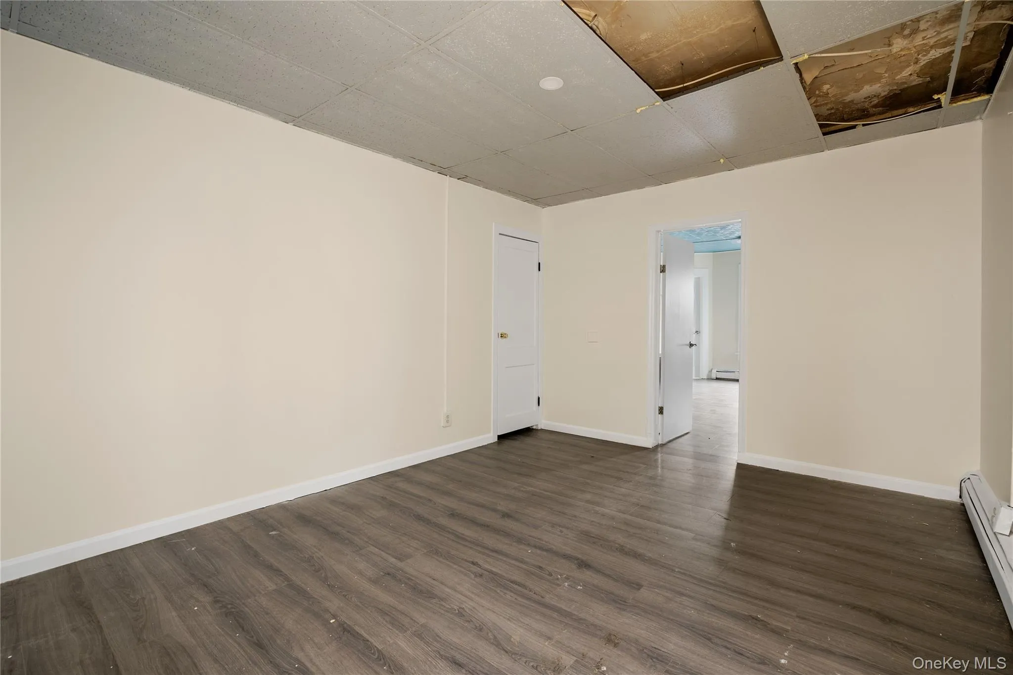 Empty room with a baseboard heating unit, dark wood-type flooring, and a drop ceiling Empty room with a baseboard heating unit, dark wood-type flooring, and a drop ceiling