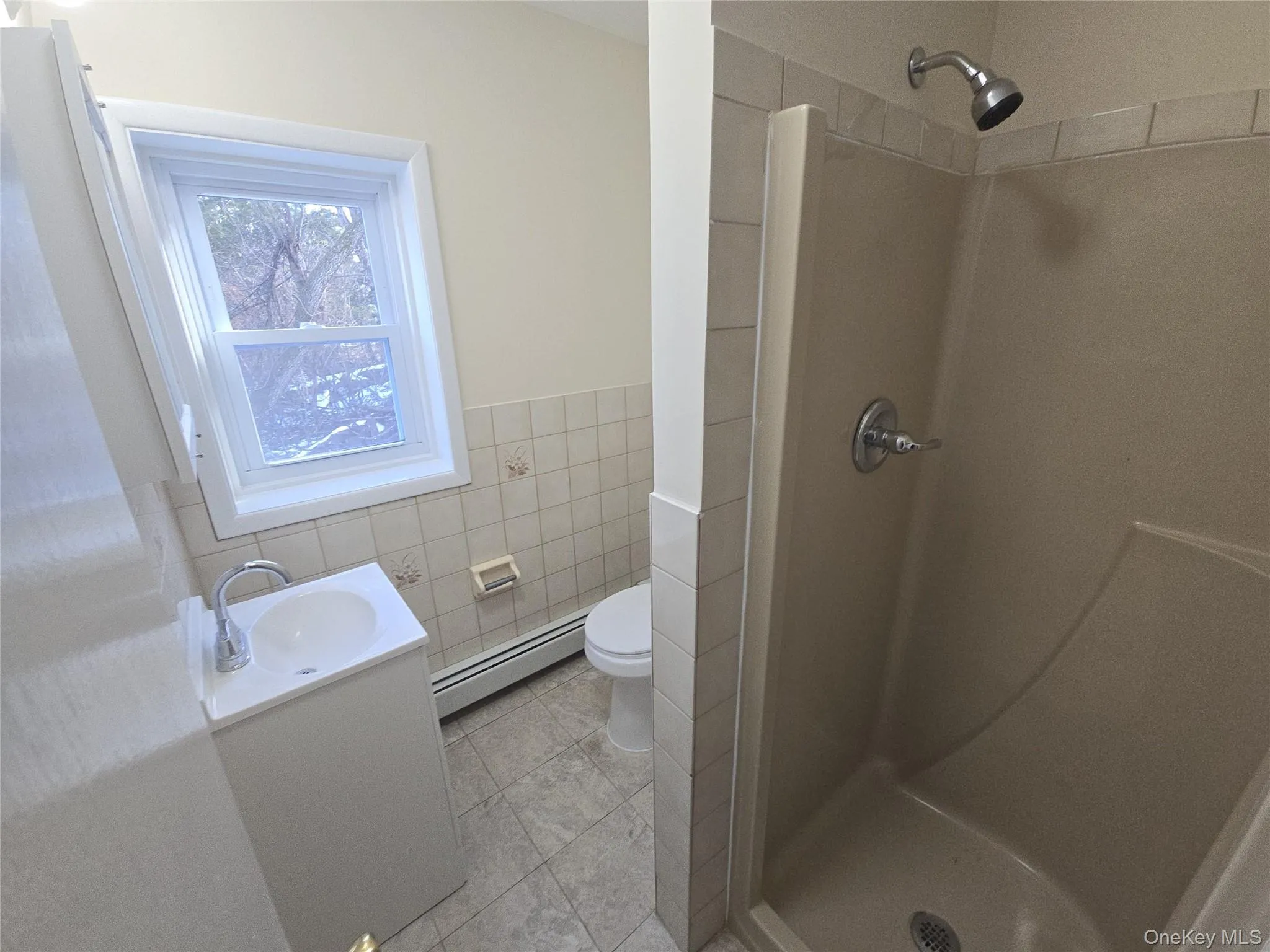 Full bath featuring vanity, tile walls, a shower stall, a baseboard heating unit, and a wainscoted wall Full bath featuring vanity, tile walls, a shower stall, a baseboard heating unit, and a wainscoted wall