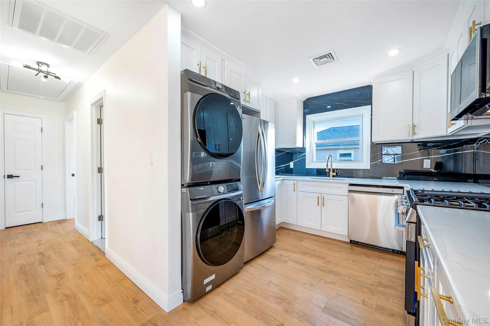 Kitchen featuring white cabinets, stainless steel appliances, backsplash, light wood finished floors, and stacked washer and clothes dryer Kitchen featuring white cabinets, stainless steel appliances, backsplash, light wood finished floors, and stacked washer and clothes dryer
