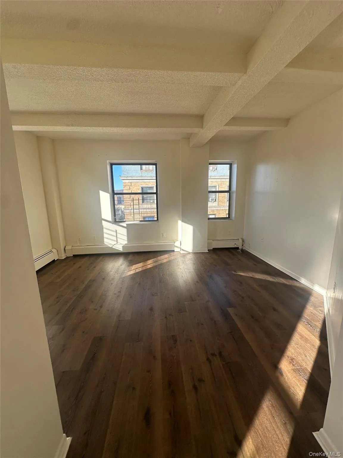 Empty room featuring beam ceiling, a textured ceiling, dark wood-type flooring, and a baseboard heating unit Empty room featuring beam ceiling, a textured ceiling, dark wood-type flooring, and a baseboard heating unit