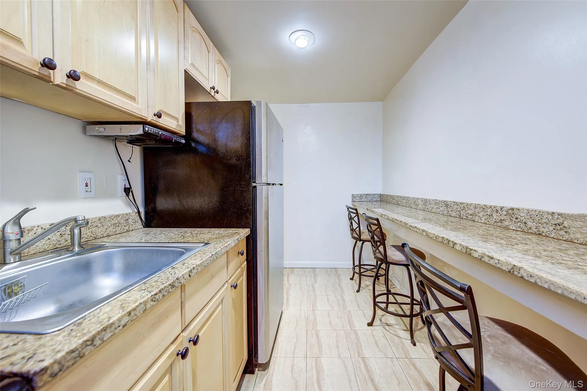 Kitchen featuring light brown cabinetry, freestanding refrigerator, light stone countertops, and a kitchen bar Kitchen featuring light brown cabinetry, freestanding refrigerator, light stone countertops, and a kitchen bar