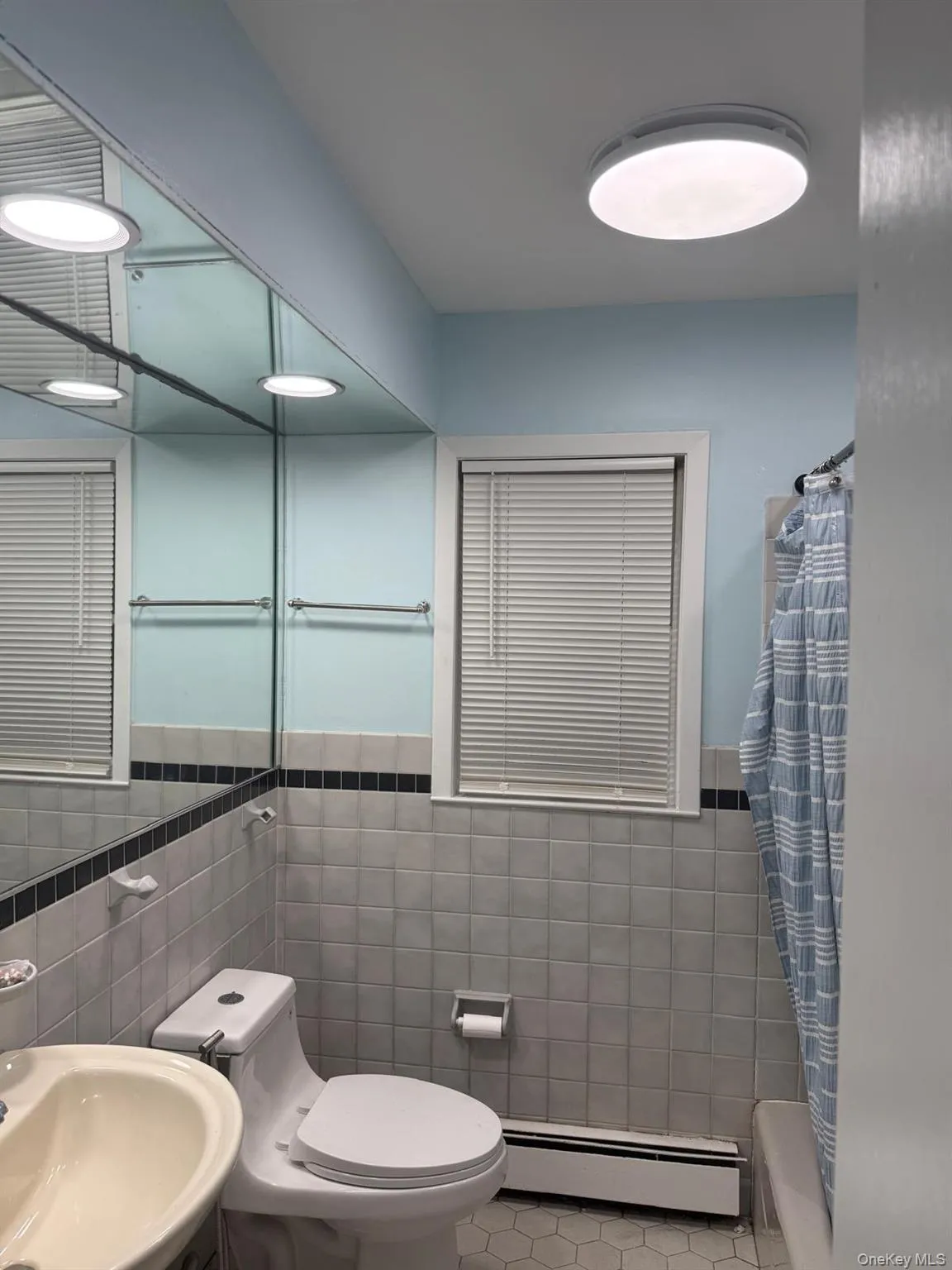 Full bathroom with tile walls, baseboard heating, shower / bath combination with curtain, wainscoting, and tile patterned floors Full bathroom with tile walls, baseboard heating, shower / bath combination with curtain, wainscoting, and tile patterned floors