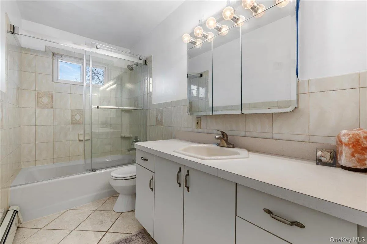 Full bath with combined bath / shower with glass door, vanity, a baseboard radiator, tile walls, and light tile patterned floors Full bath with combined bath / shower with glass door, vanity, a baseboard radiator, tile walls, and light tile patterned floors