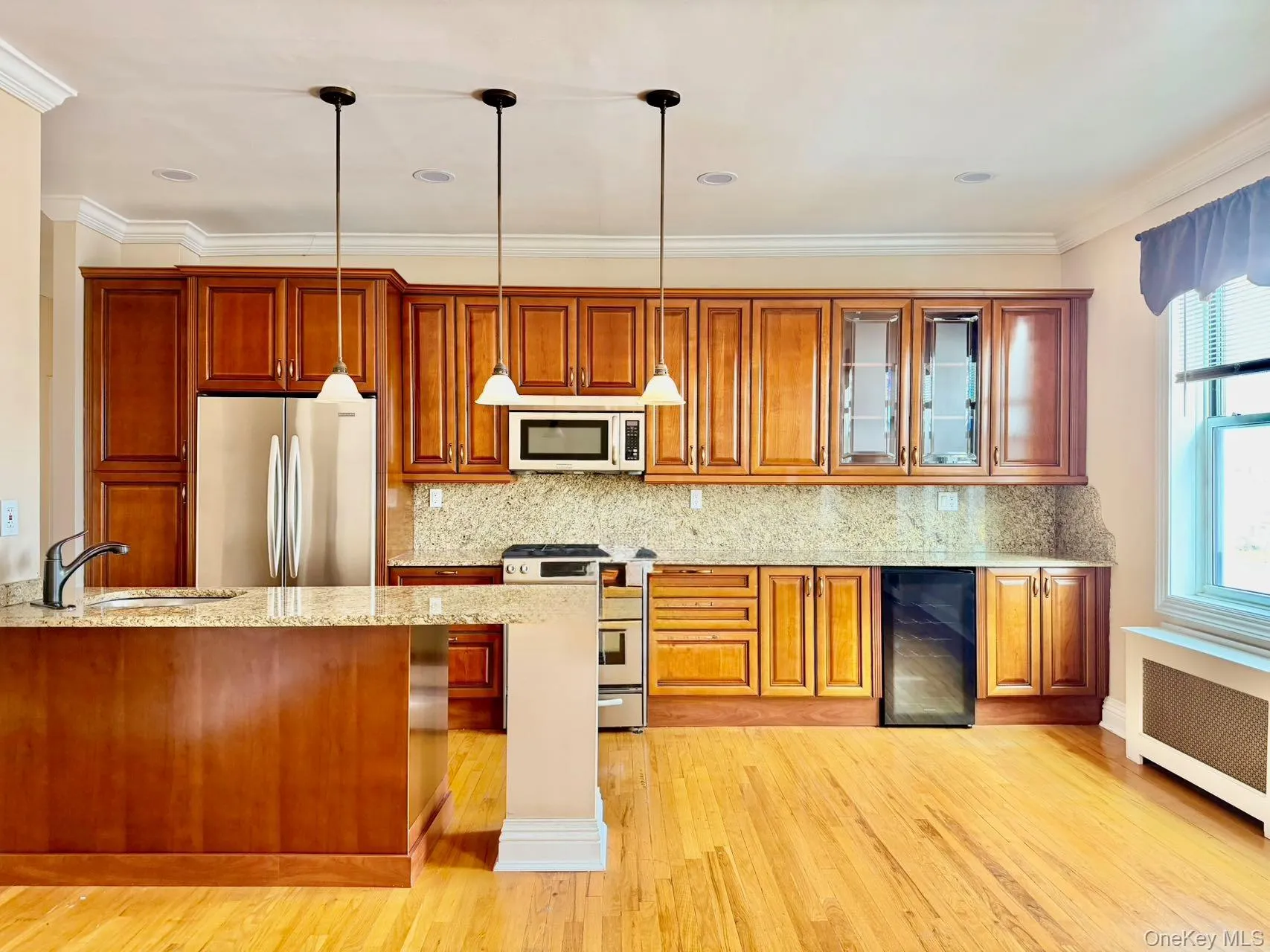 Kitchen with brown cabinetry, appliances with stainless steel finishes, light stone countertops, decorative light fixtures, and wine cooler Kitchen with brown cabinetry, appliances with stainless steel finishes, light stone countertops, decorative light fixtures, and wine cooler