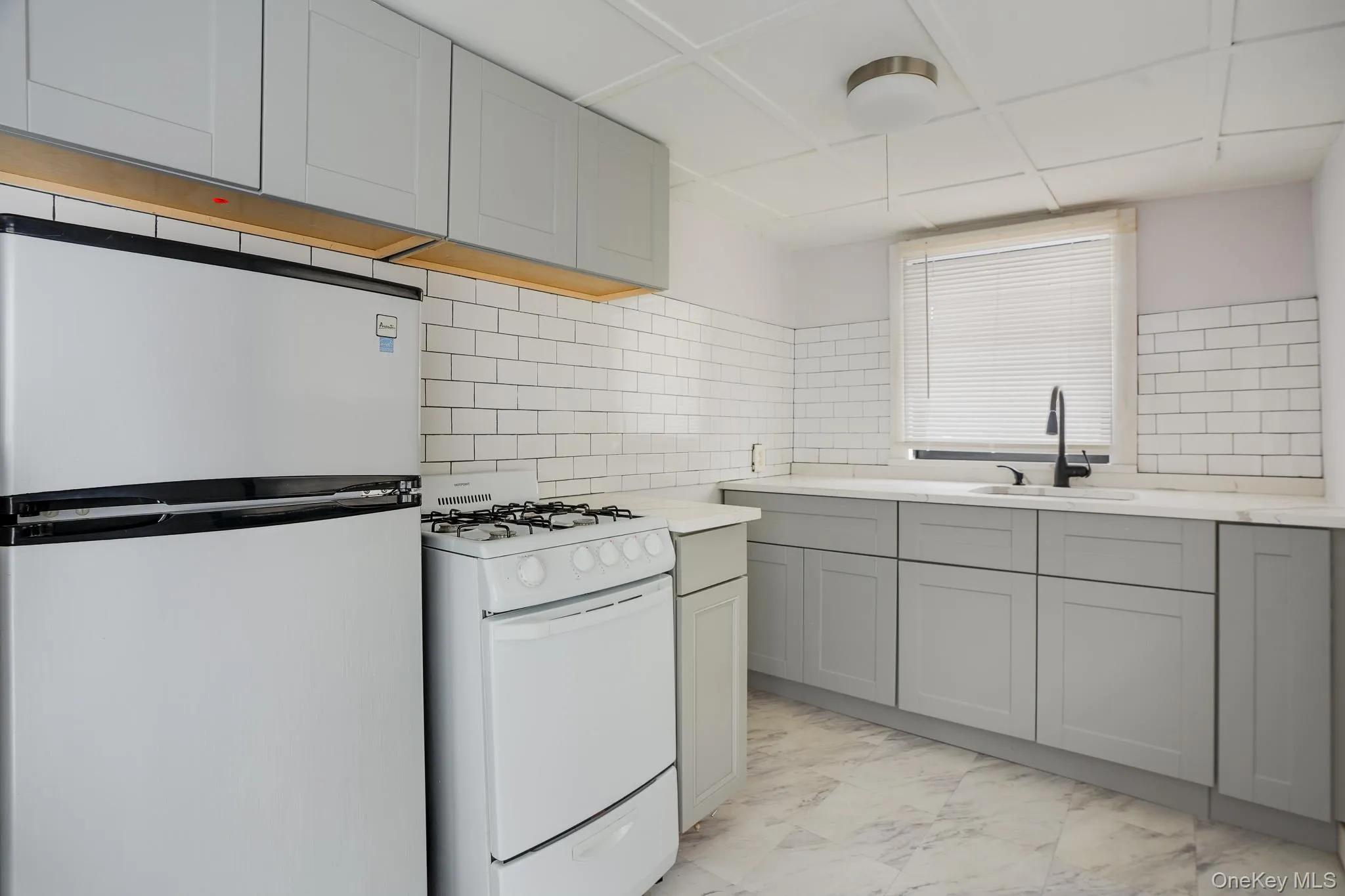 Kitchen with white appliances, gray cabinetry, light marble finish flooring, and decorative backsplash Kitchen with white appliances, gray cabinetry, light marble finish flooring, and decorative backsplash