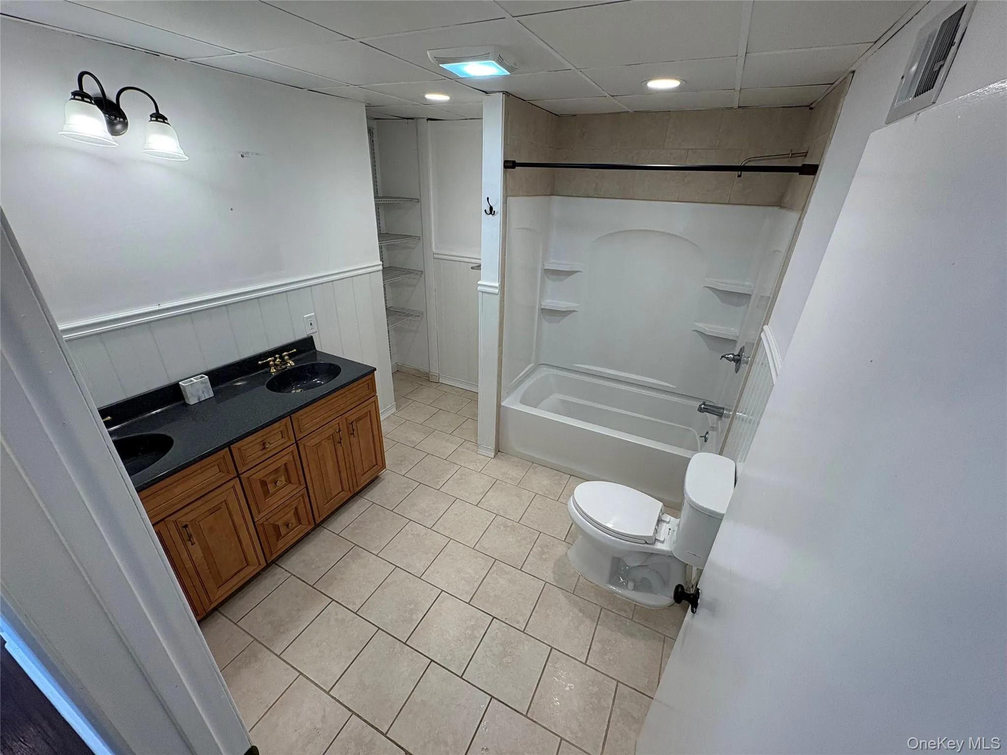 Full bath with double vanity, light tile patterned floors, a paneled ceiling, wainscoting, and tub / shower combination Full bath with double vanity, light tile patterned floors, a paneled ceiling, wainscoting, and tub / shower combination