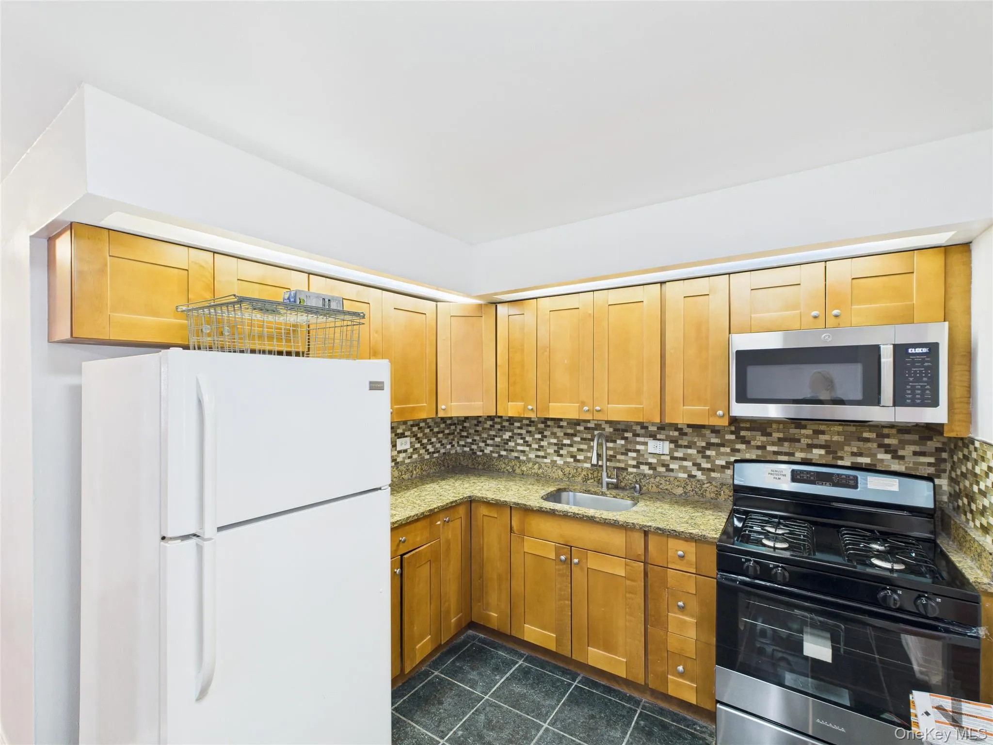 Kitchen with appliances with stainless steel finishes, light stone countertops, backsplash, brown cabinetry, and dark tile patterned flooring Kitchen with appliances with stainless steel finishes, light stone countertops, backsplash, brown cabinetry, and dark tile patterned flooring