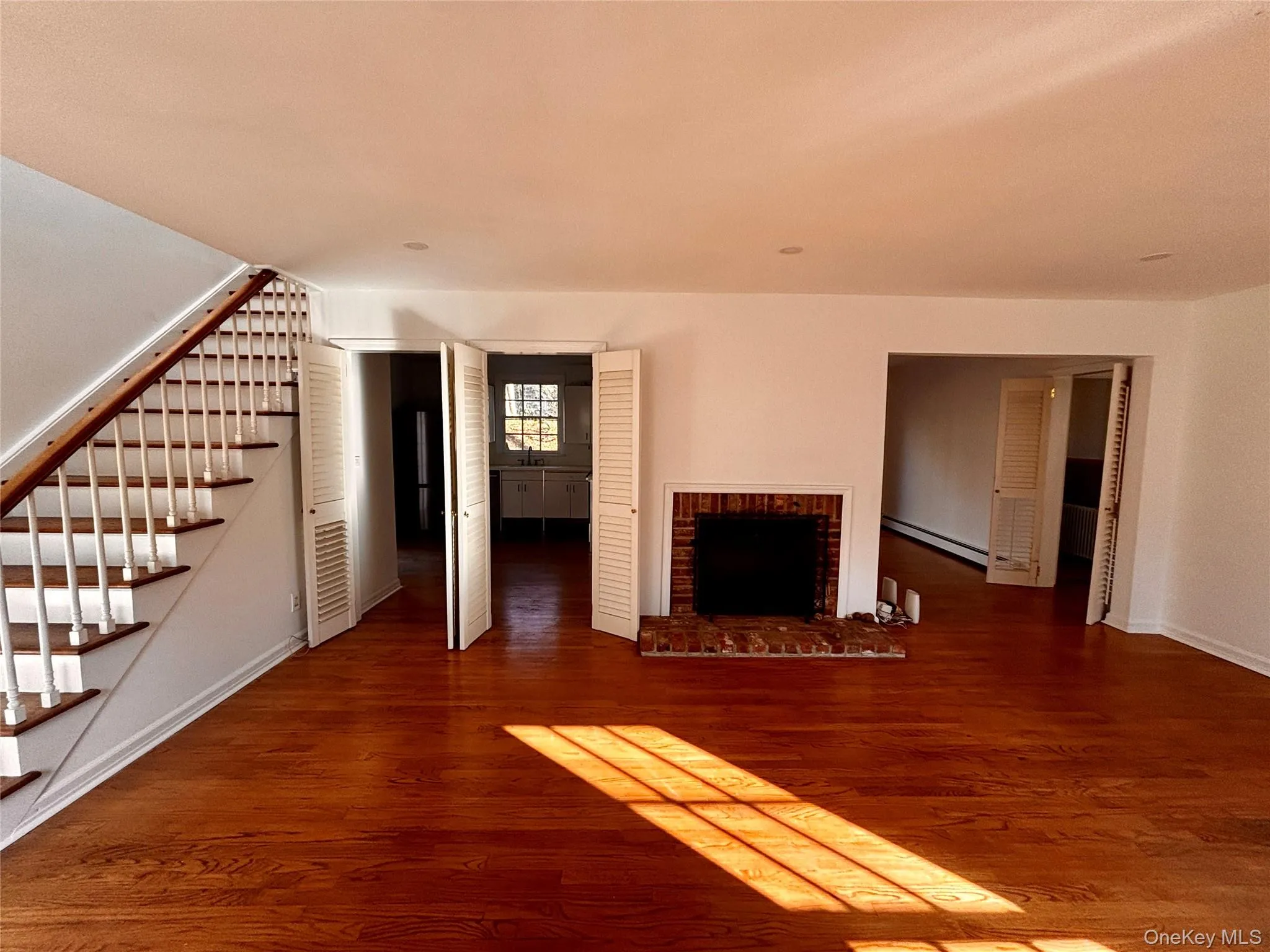 Unfurnished living room featuring stairs, a brick fireplace, dark wood-style floors, and baseboard heating Unfurnished living room featuring stairs, a brick fireplace, dark wood-style floors, and baseboard heating