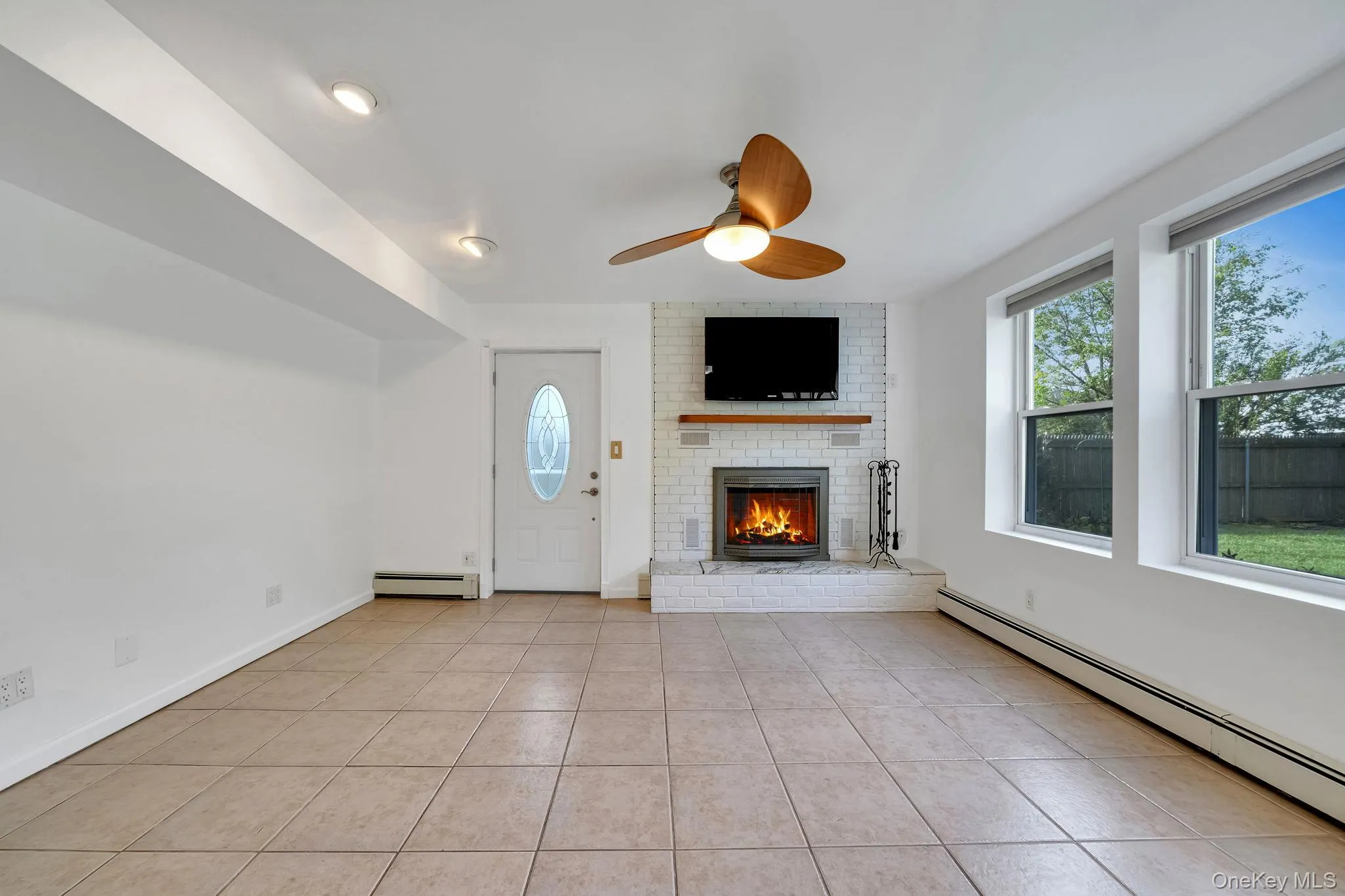 Unfurnished living room with a baseboard radiator, light tile patterned floors, a brick fireplace, a ceiling fan, and healthy amount of natural light Unfurnished living room with a baseboard radiator, light tile patterned floors, a brick fireplace, a ceiling fan, and healthy amount of natural light
