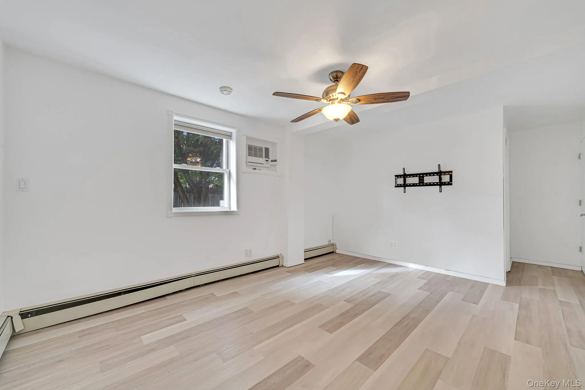 Empty room featuring light wood-style flooring, baseboard heating, ceiling fan, and a wall mounted air conditioner Empty room featuring light wood-style flooring, baseboard heating, ceiling fan, and a wall mounted air conditioner