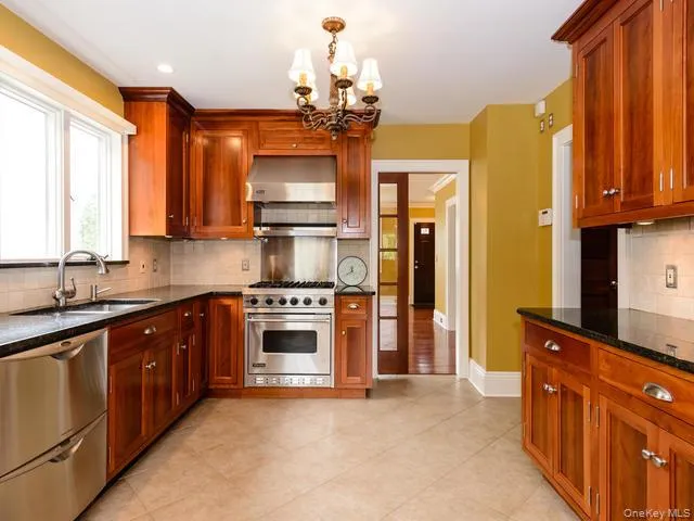 Kitchen with tasteful backsplash, appliances with stainless steel finishes, a chandelier, wall chimney exhaust hood, and dark stone countertops Kitchen with tasteful backsplash, appliances with stainless steel finishes, a chandelier, wall chimney exhaust hood, and dark stone countertops