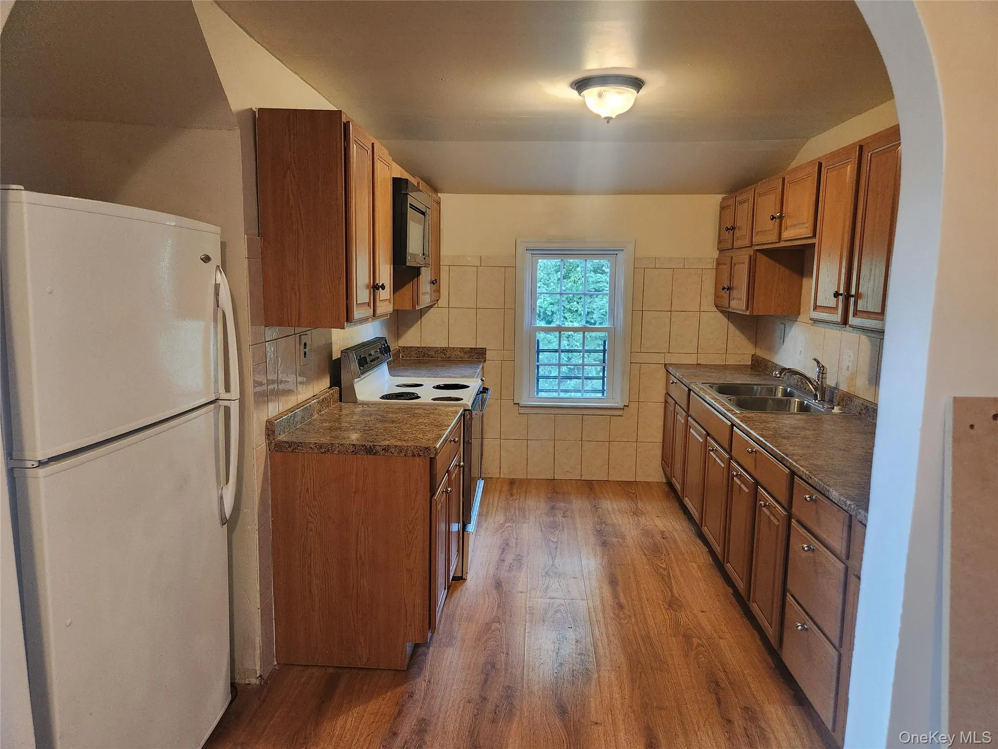 Kitchen with white appliances, dark countertops, arched walkways, brown cabinets, and tile walls Kitchen with white appliances, dark countertops, arched walkways, brown cabinets, and tile walls