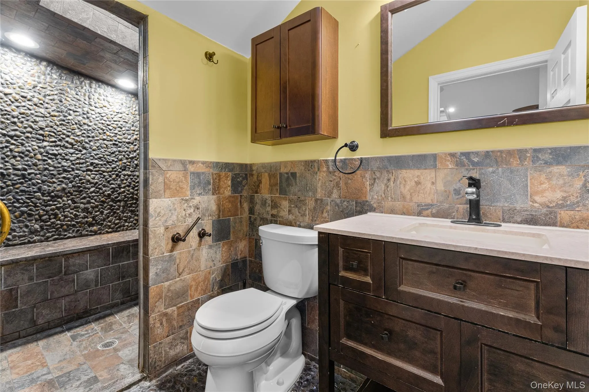 Bathroom with tile walls, a wainscoted wall, vanity, tiled shower, and stone tile flooring Bathroom with tile walls, a wainscoted wall, vanity, tiled shower, and stone tile flooring