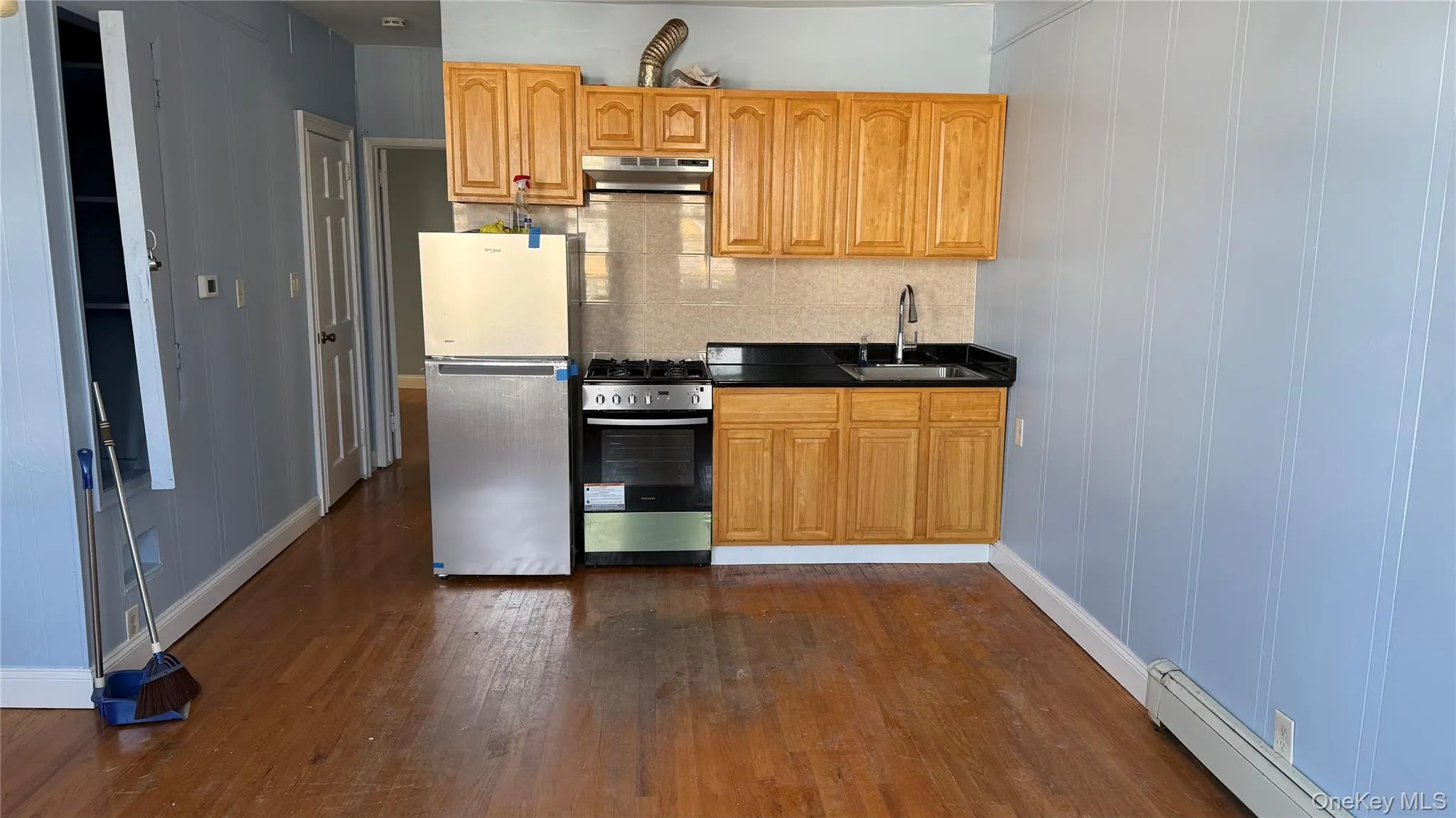Kitchen featuring baseboard heating, appliances with stainless steel finishes, dark countertops, tasteful backsplash, and under cabinet range hood Kitchen featuring baseboard heating, appliances with stainless steel finishes, dark countertops, tasteful backsplash, and under cabinet range hood
