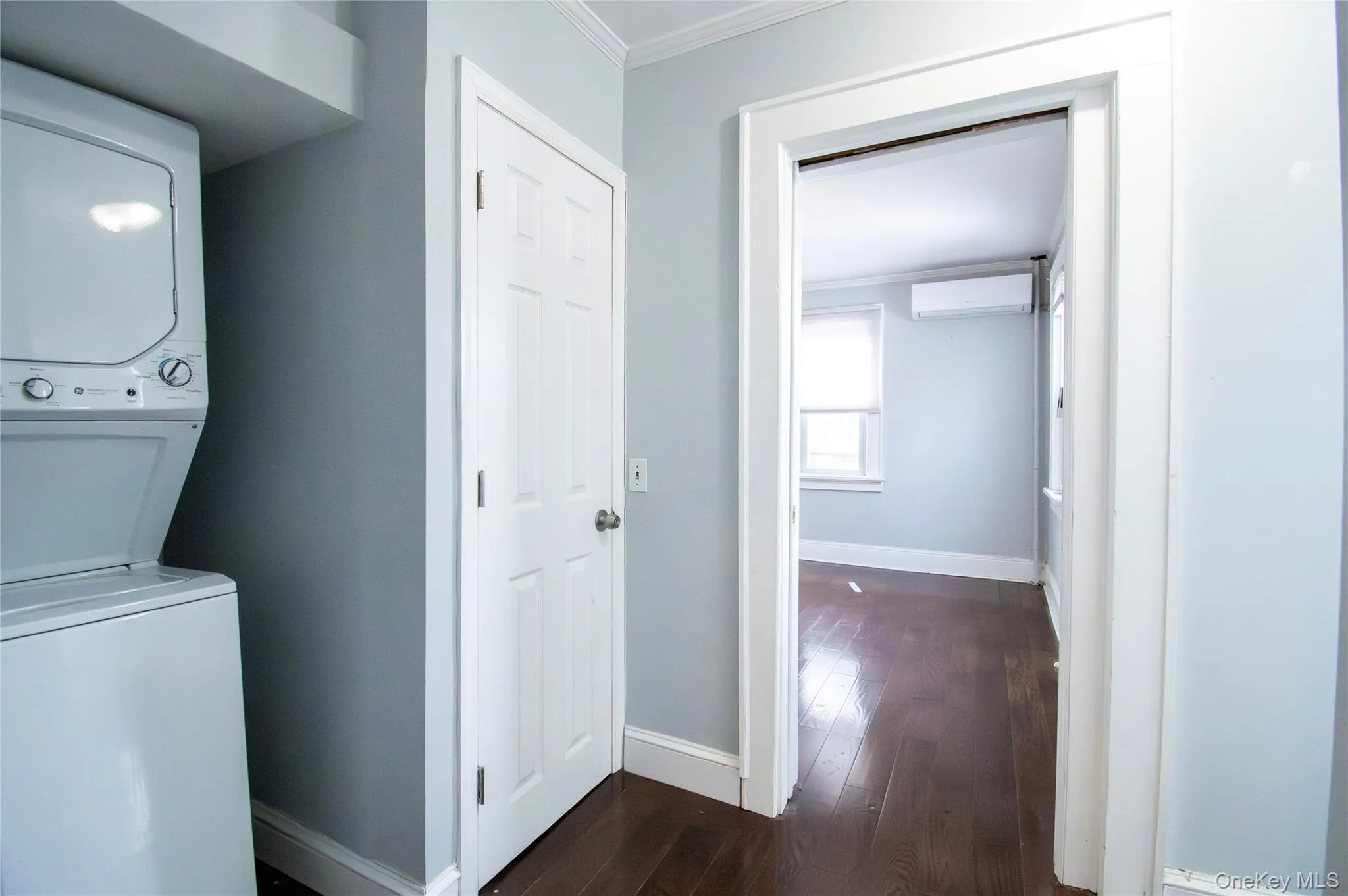 Washroom with crown molding, dark wood-style flooring, stacked washer / drying machine, and a wall unit AC Washroom with crown molding, dark wood-style flooring, stacked washer / drying machine, and a wall unit AC