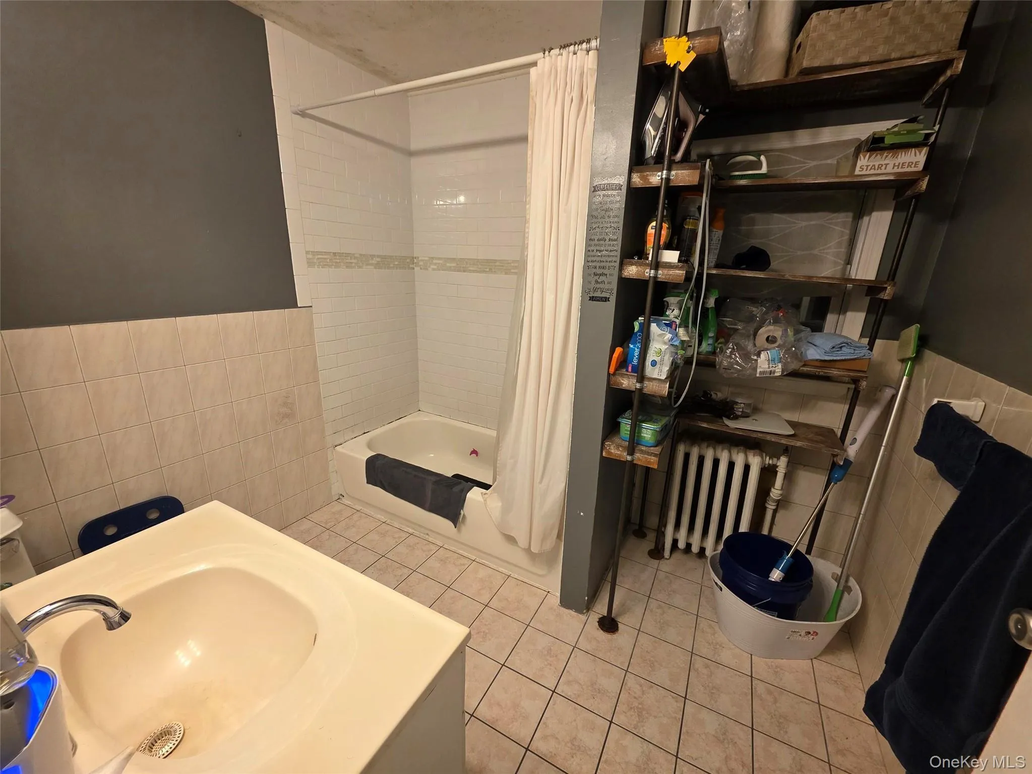 Full bath featuring tile walls, shower / bath combo, vanity, radiator heating unit, and light tile patterned floors Full bath featuring tile walls, shower / bath combo, vanity, radiator heating unit, and light tile patterned floors