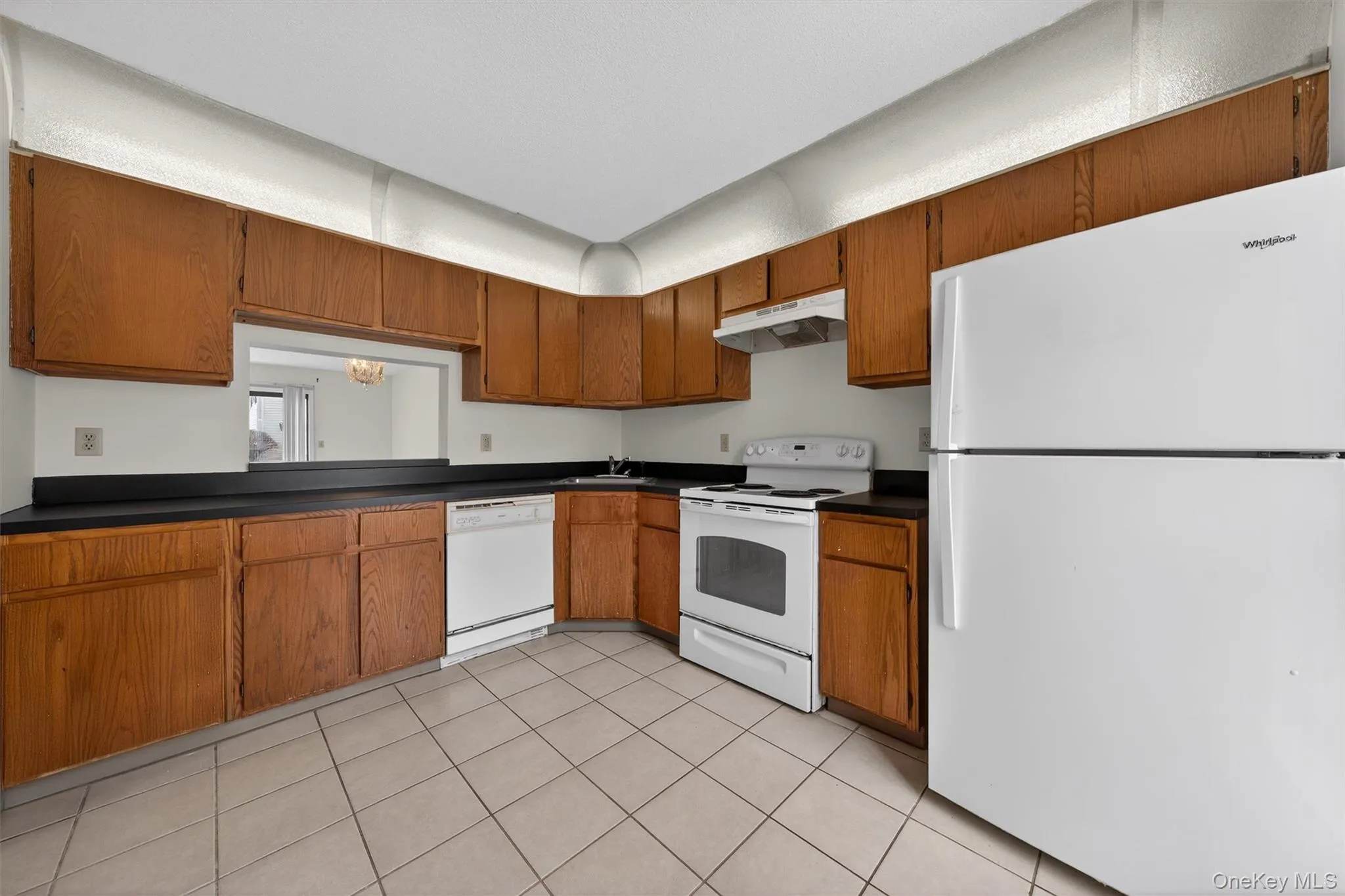 Kitchen featuring white appliances, brown cabinets, dark countertops, under cabinet range hood, and light tile patterned flooring Kitchen featuring white appliances, brown cabinets, dark countertops, under cabinet range hood, and light tile patterned flooring