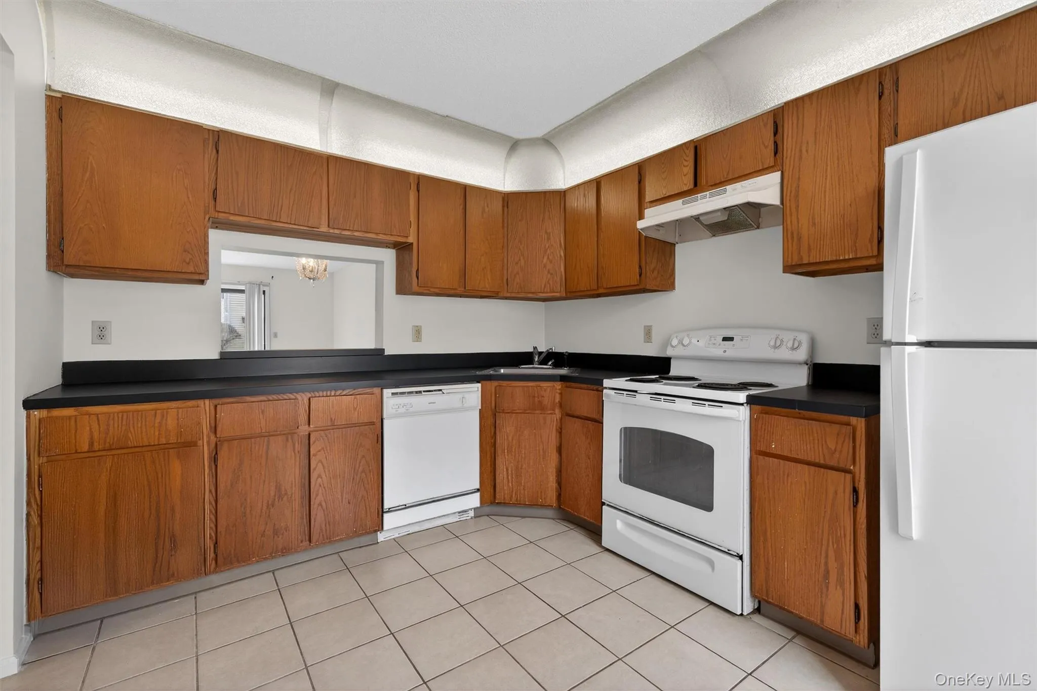 Kitchen with white appliances, brown cabinets, under cabinet range hood, dark countertops, and light tile patterned floors Kitchen with white appliances, brown cabinets, under cabinet range hood, dark countertops, and light tile patterned floors