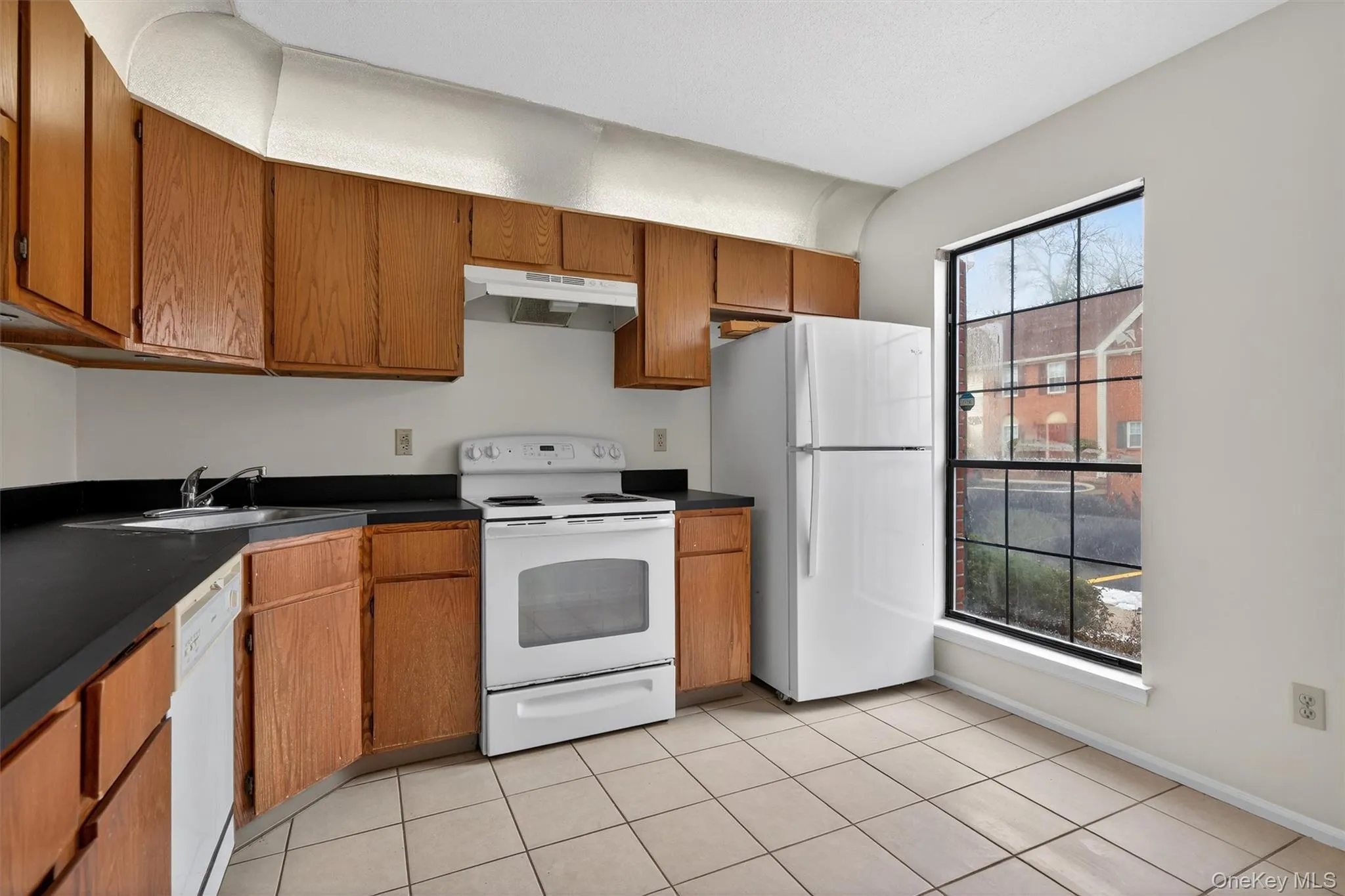 Kitchen featuring white appliances, dark countertops, brown cabinetry, under cabinet range hood, and light tile patterned floors Kitchen featuring white appliances, dark countertops, brown cabinetry, under cabinet range hood, and light tile patterned floors