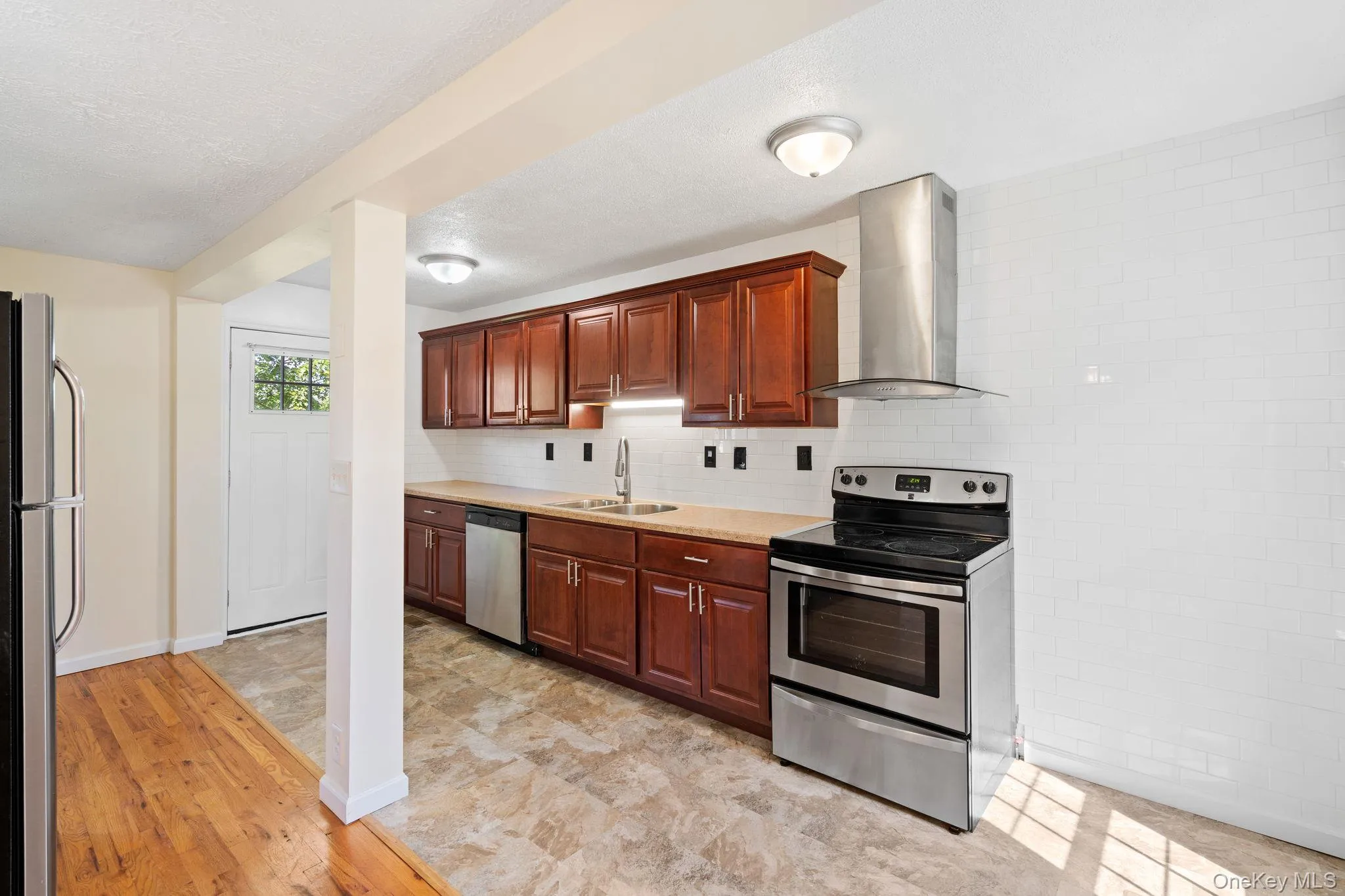 Kitchen featuring appliances with stainless steel finishes, wall chimney exhaust hood, decorative backsplash, light wood-type flooring, and a textured ceiling Kitchen featuring appliances with stainless steel finishes, wall chimney exhaust hood, decorative backsplash, light wood-type flooring, and a textured ceiling
