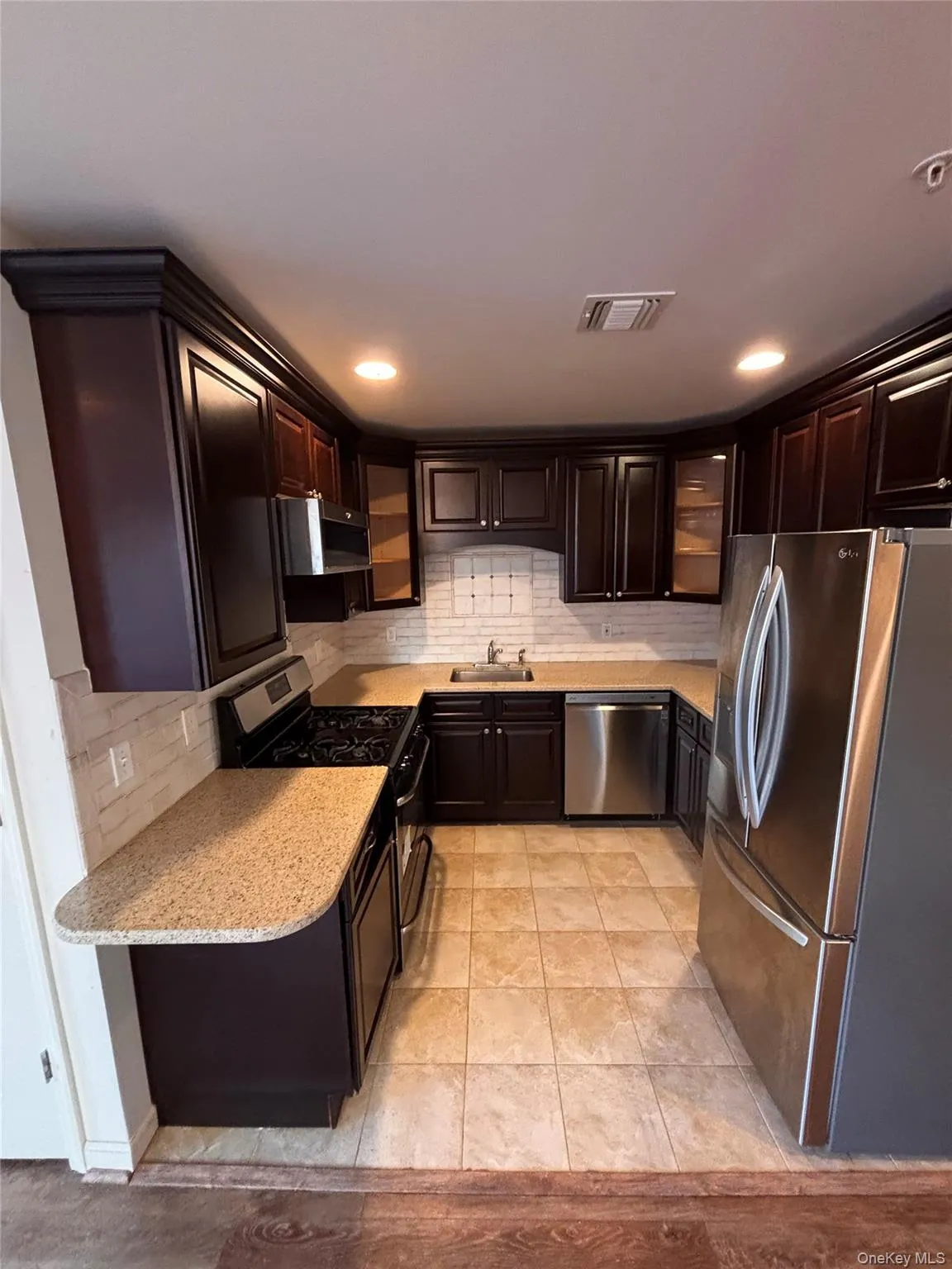 Kitchen with decorative backsplash, appliances with stainless steel finishes, glass insert cabinets, light stone counters, and light tile patterned floors Kitchen with decorative backsplash, appliances with stainless steel finishes, glass insert cabinets, light stone counters, and light tile patterned floors