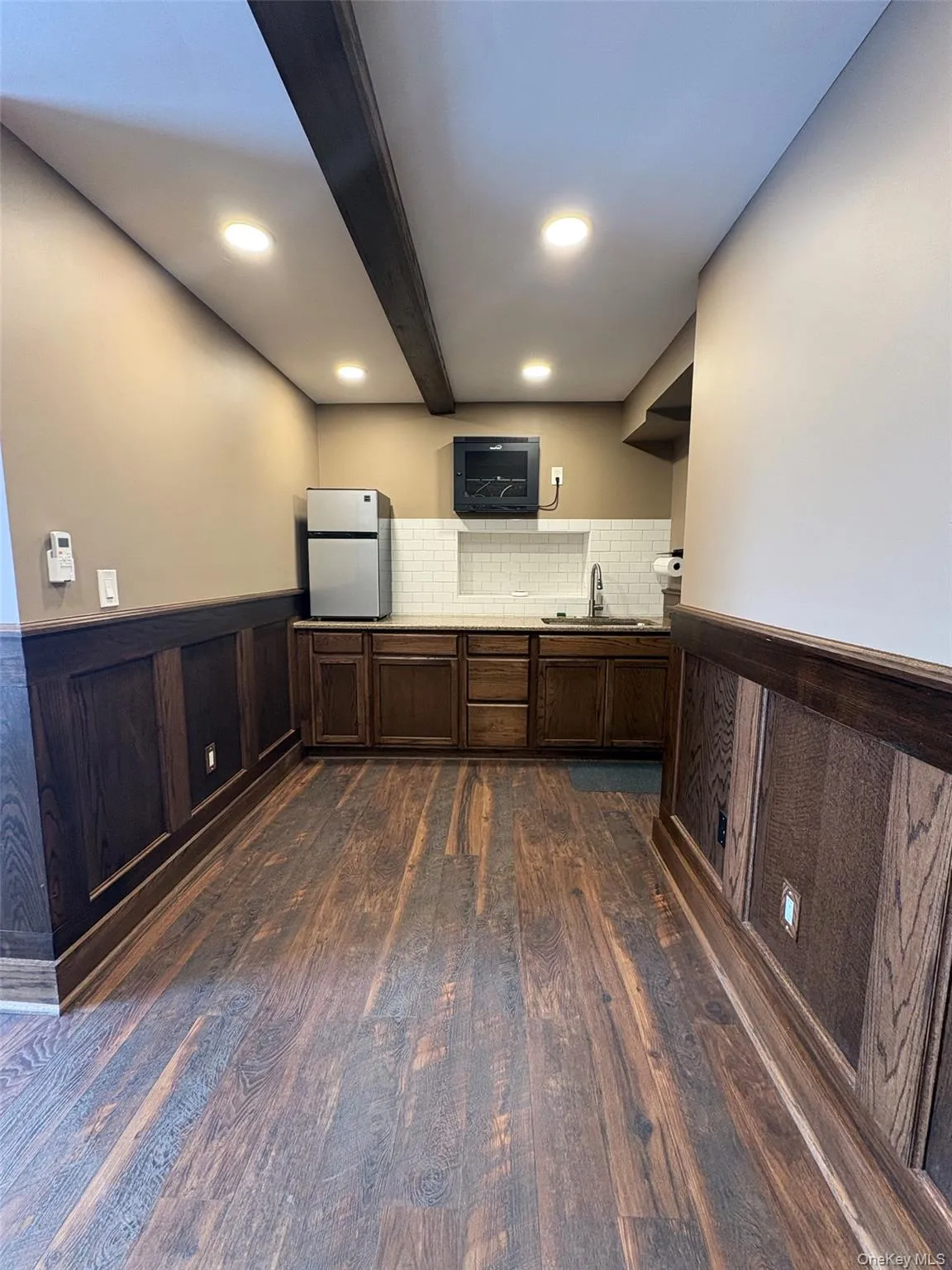 Bar area featuring a wainscoted wall, freestanding refrigerator, dark brown cabinets, dark wood-style flooring, and light countertops Bar area featuring a wainscoted wall, freestanding refrigerator, dark brown cabinets, dark wood-style flooring, and light countertops
