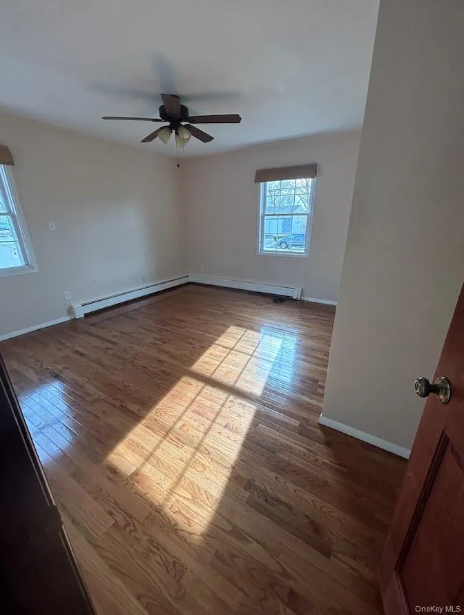 Empty room featuring dark wood-style flooring, plenty of natural light, ceiling fan, and baseboard heating Empty room featuring dark wood-style flooring, plenty of natural light, ceiling fan, and baseboard heating