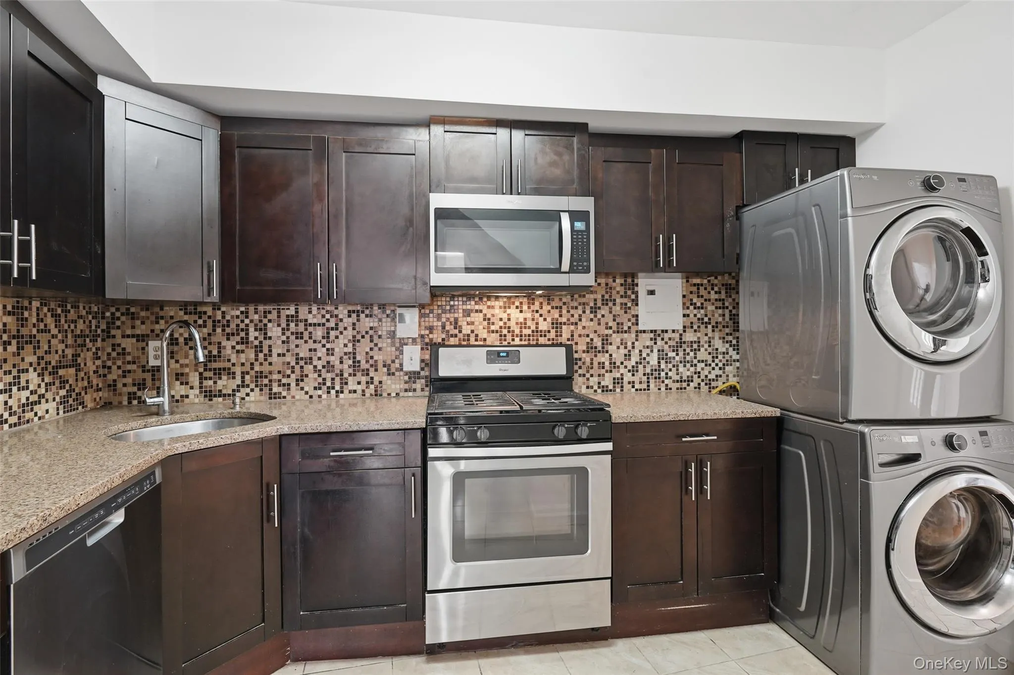 Kitchen featuring dark brown cabinets, appliances with stainless steel finishes, light stone counters, and estacked washer and dryer Kitchen featuring dark brown cabinets, appliances with stainless steel finishes, light stone counters, and estacked washer and dryer