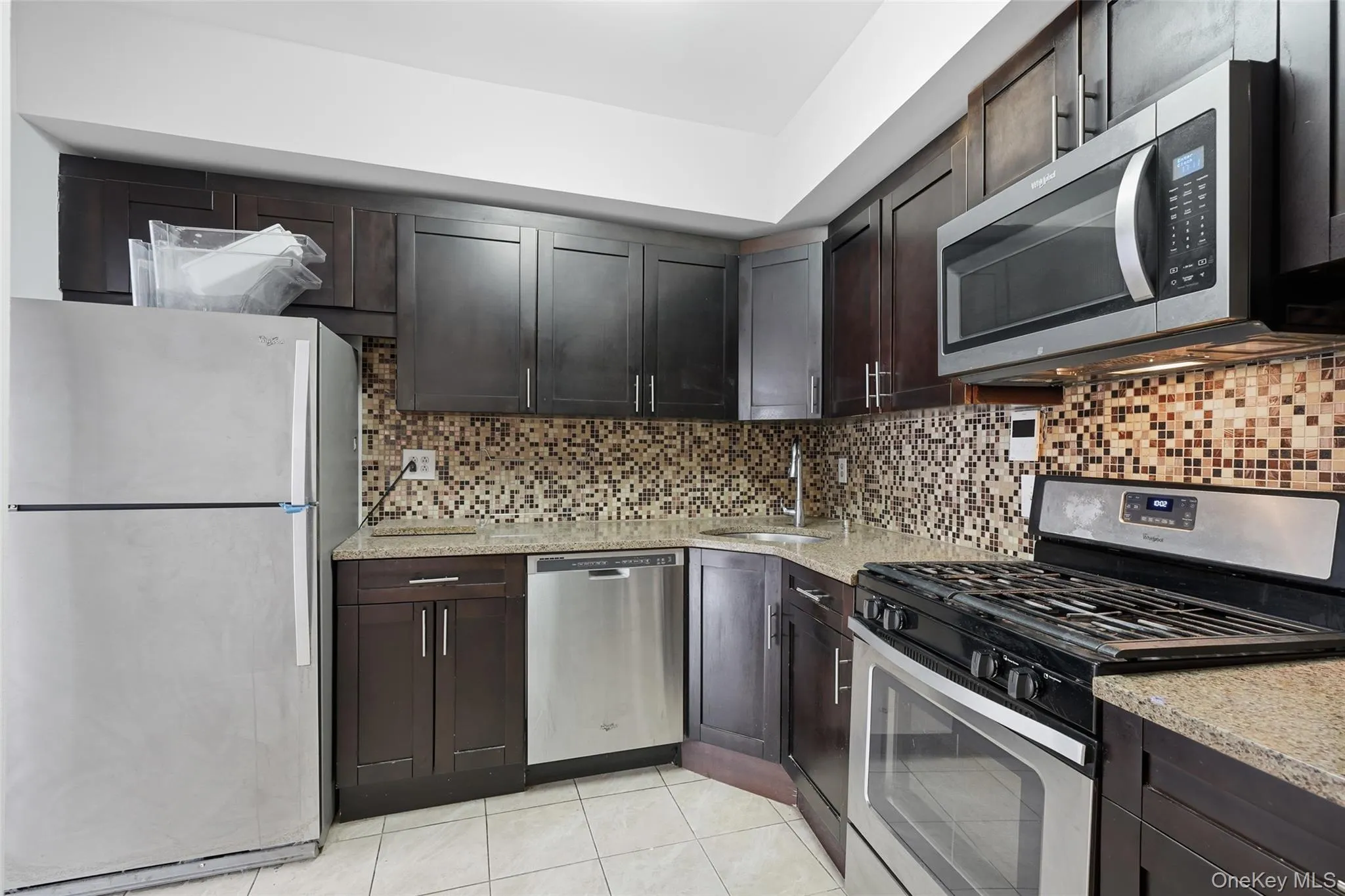 Kitchen featuring appliances with stainless steel finishes, dark brown cabinetry, light stone countertops, light tile patterned floors, and backsplash Kitchen featuring appliances with stainless steel finishes, dark brown cabinetry, light stone countertops, light tile patterned floors, and backsplash