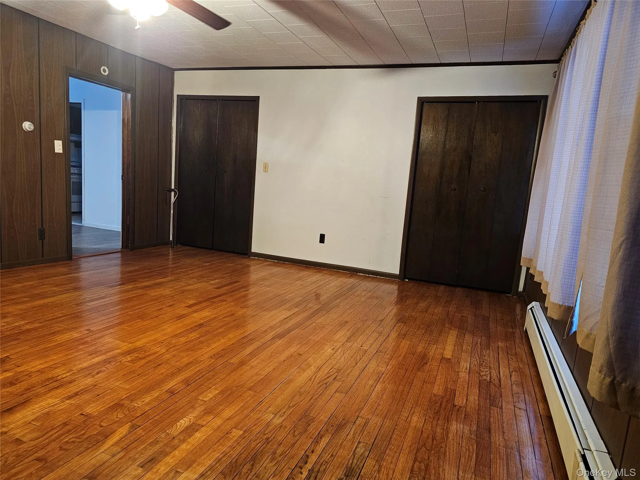 Unfurnished bedroom featuring a baseboard heating unit, two closets, hardwood / wood-style floors, ceiling fan, and wooden walls Unfurnished bedroom featuring a baseboard heating unit, two closets, hardwood / wood-style floors, ceiling fan, and wooden walls