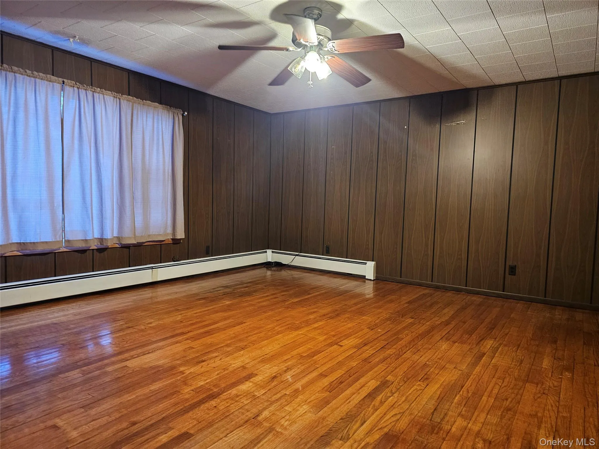 Unfurnished room featuring dark wood-type flooring, a baseboard radiator, wooden walls, and a ceiling fan Unfurnished room featuring dark wood-type flooring, a baseboard radiator, wooden walls, and a ceiling fan