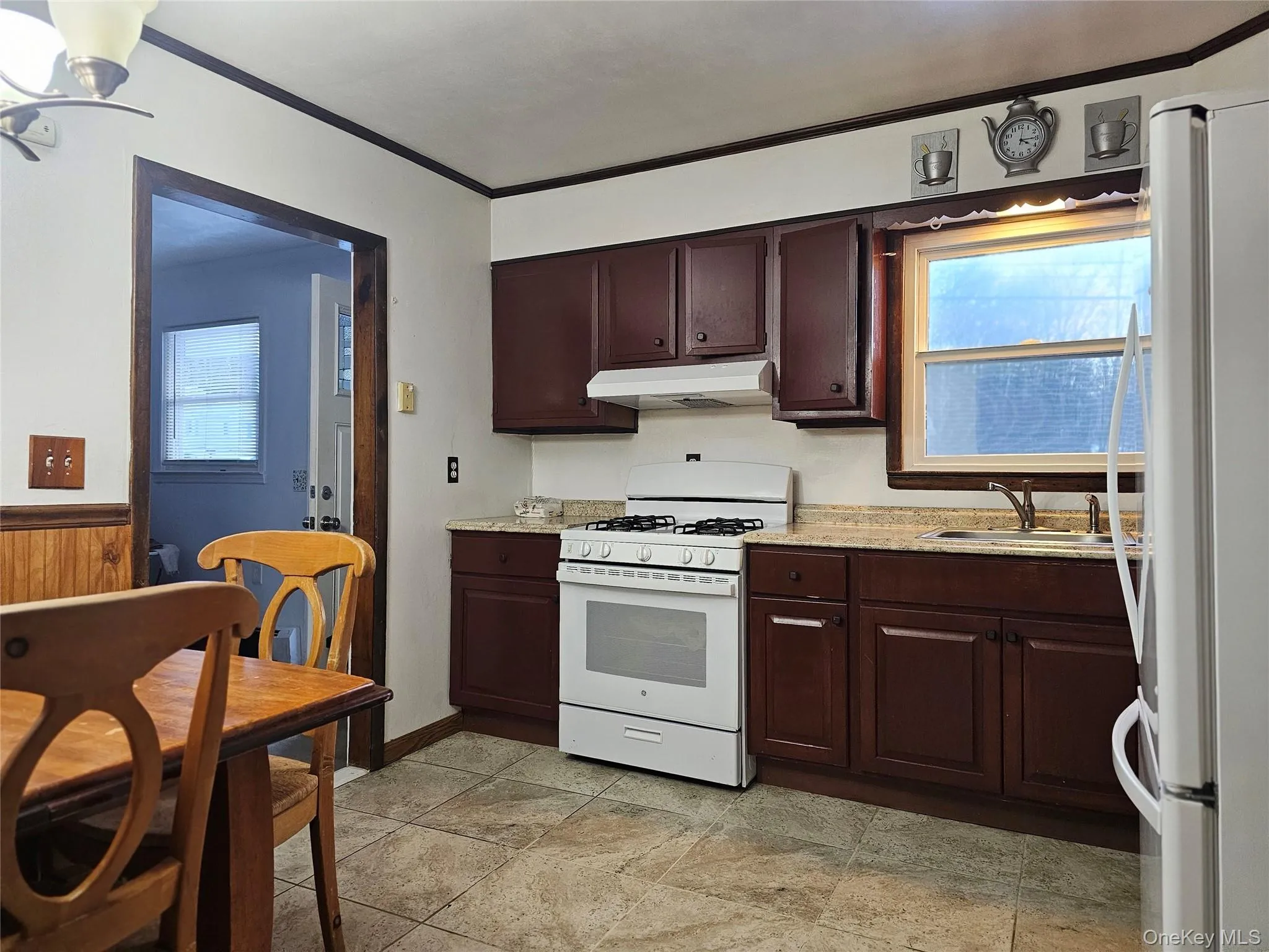 Kitchen with white appliances, dark brown cabinets, under cabinet range hood, ornamental molding, and light stone countertops Kitchen with white appliances, dark brown cabinets, under cabinet range hood, ornamental molding, and light stone countertops