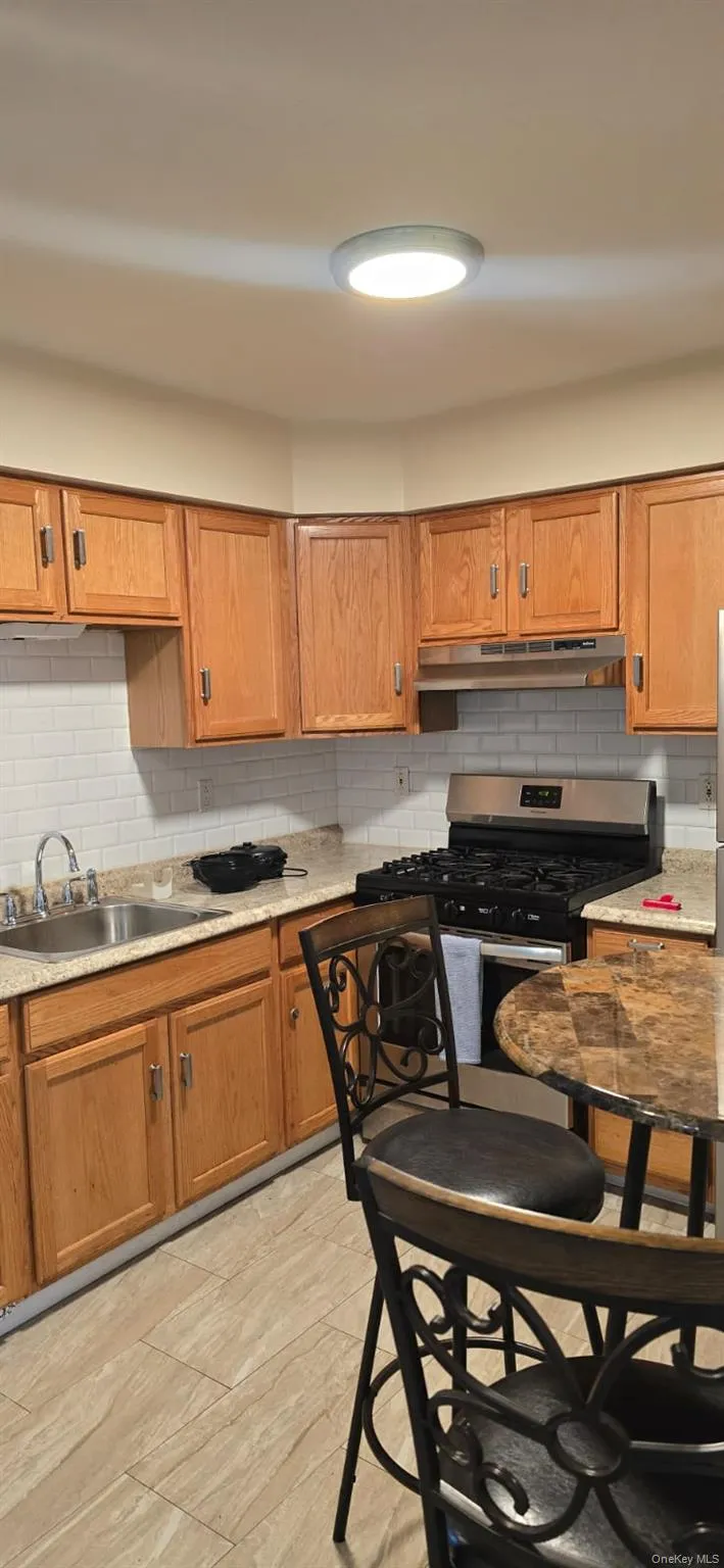 Kitchen with backsplash, gas range, brown cabinets, under cabinet range hood, and light stone countertops Kitchen with backsplash, gas range, brown cabinets, under cabinet range hood, and light stone countertops