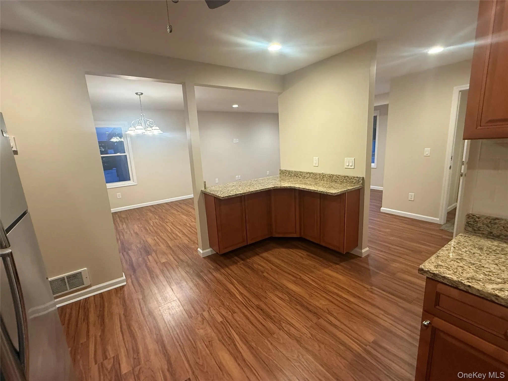Kitchen featuring brown cabinetry, light stone counters, dark wood-type flooring, freestanding refrigerator, and pendant lighting Kitchen featuring brown cabinetry, light stone counters, dark wood-type flooring, freestanding refrigerator, and pendant lighting